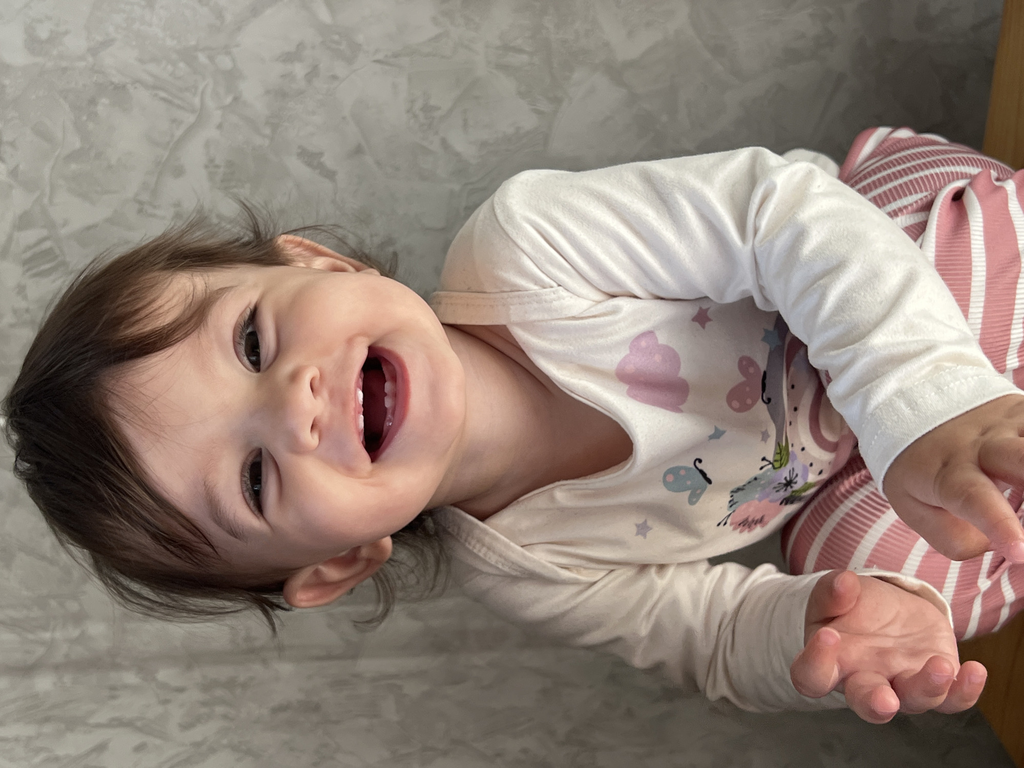 A close-up, slightly angled view shows a young child lying on their side on a surface with a grey, textured background. The child is fair-skinned with dark hair and dark eyes. They are smiling broadly, revealing their teeth. The child is wearing a light-colored long-sleeved shirt with a print of clouds, butterflies, and rainbows. They also have striped pink and white pants. The child's hands are positioned towards the viewer. There is a wooden edge visible behind their legs. The location of Elena, Bulgaria, is not apparent in this image.