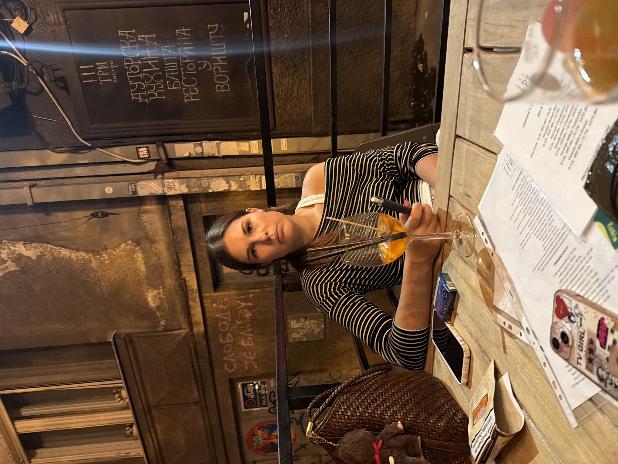 A woman is seated at a wooden table outdoors in front of a building with a dark door and graffiti on the wall. She is wearing a black and white striped off-the-shoulder top and is looking towards the camera with a neutral expression. In front of her is a glass of orange liquid with straws and a vape pen. On the table are a smartphone, a pack of cigarettes, a brown woven bag with a small bear keychain attached, and some papers. The background shows a building with windows and what appears to be a menu or signage written in Cyrillic script. The setting suggests an outdoor seating area of a restaurant or bar in Belgrade, Serbia. The graffiti on the wall includes Cyrillic text.
