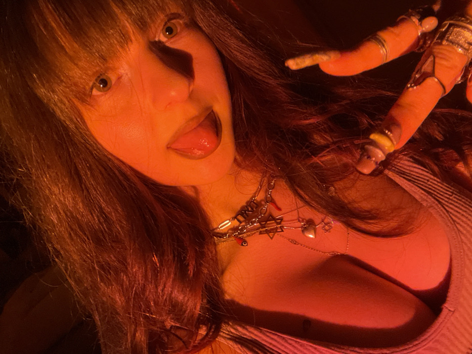 A woman with bangs sticks her tongue out. Her hair is reddish brown and her eyes are green. She wears multiple necklaces with various charms. Her cleavage is exposed, and she wears a gray tank top. A hand with multiple rings is raised near her head, appearing to hold a rolled cigarette or joint. The image has a reddish-orange tint.