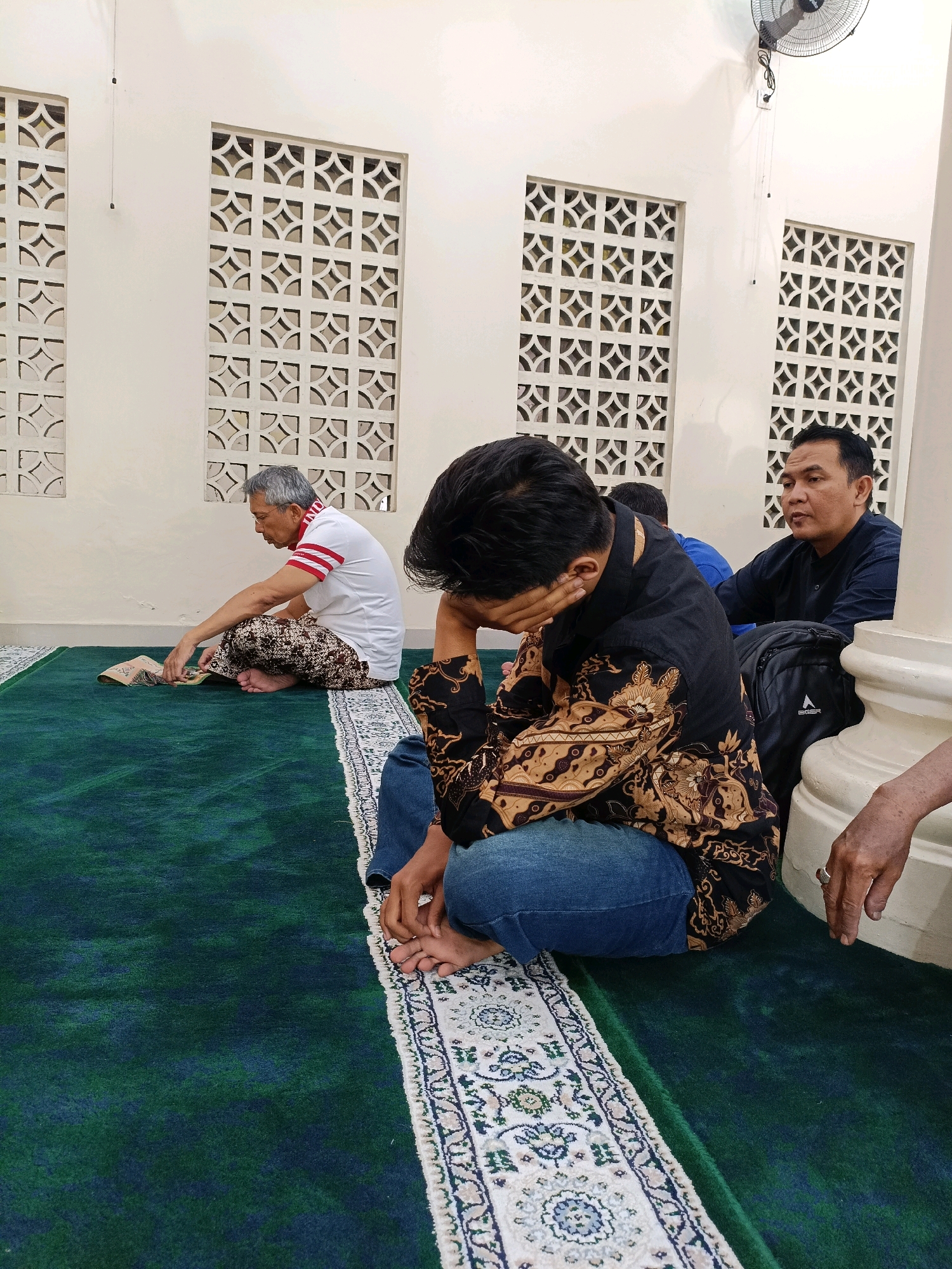 The image depicts an indoor scene, likely a prayer room or mosque, given the green carpet and the individuals' attire and seated positions. Three men are visible. In the foreground, a man in a black shirt with a gold and brown patterned design and blue jeans is seated, with his head bowed and hand over his face, suggesting distress or contemplation. To his left, an older man with grey hair, wearing a white polo shirt with red stripes and patterned pants, is seated on the carpet, looking down at an open book or Quran. To the right, another man in a dark blue shirt is seated and looking towards the left, with a backpack beside him. The walls are white with decorative cut-out panels. A ceiling fan is visible in the upper right corner. The floor is covered with a green carpet and a decorative rug with a white border featuring a blue and floral pattern. The scene appears to be in Maguwoharjo, Indonesia, a location known for its religious sites.