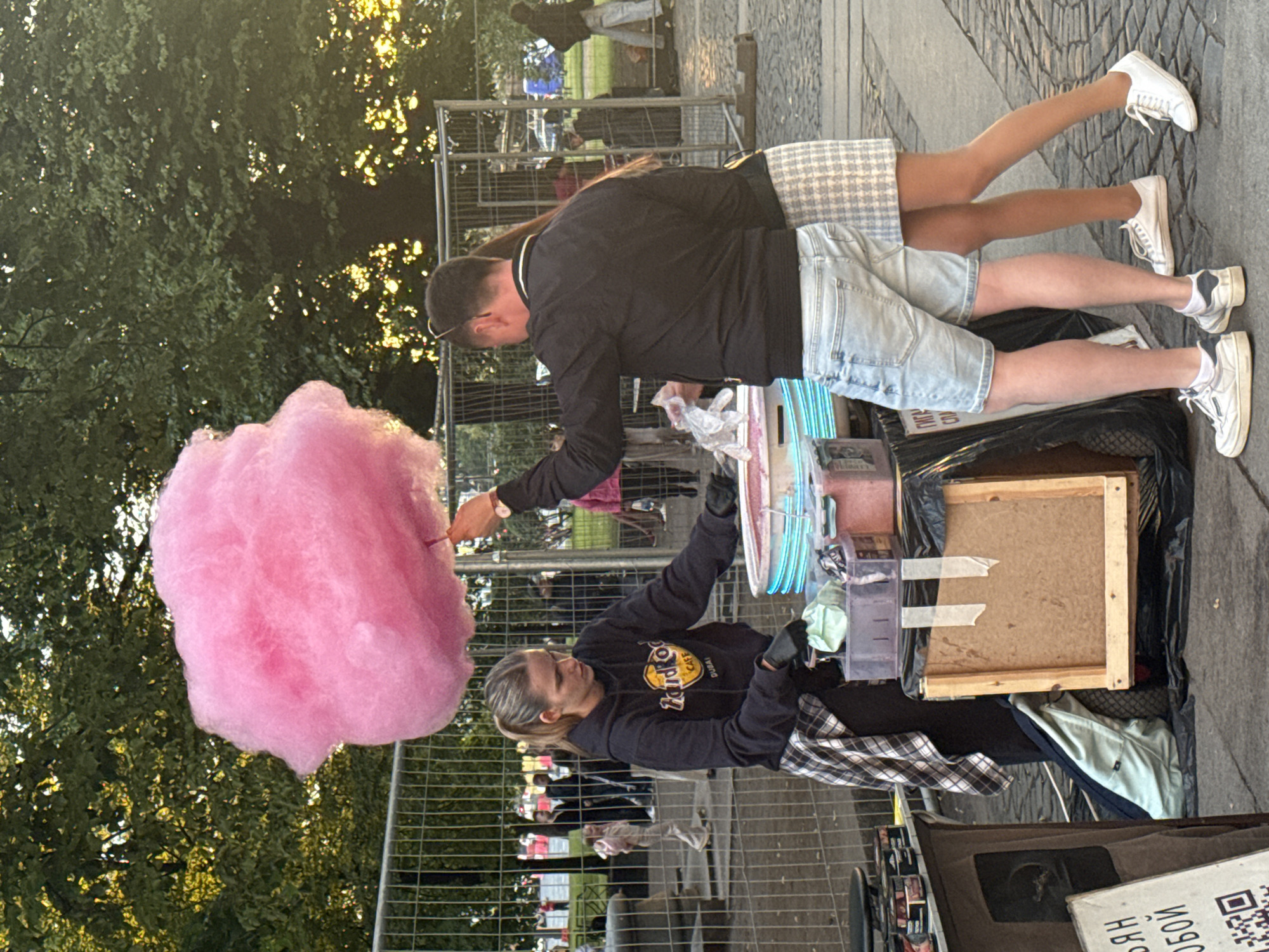 The image shows a street scene where a woman is making cotton candy. She is wearing a dark sweatshirt with a logo that says "Hard Rock Cafe" and a plaid shirt. She is holding a cone and appears to be handing it to a person off-camera. The cotton candy is large and pink. A man is standing next to her, wearing a black jacket and light-colored shorts, and also appears to be involved in the transaction. He is holding a bag. In the background, there are metal fences and trees with sunlight filtering through the leaves, suggesting it is daytime, possibly late afternoon or early evening. The ground is paved with cobblestones. Further in the background, other people and a fence are visible, hinting at a public gathering or event. There is some text visible on a white box in the foreground, but it is partially obscured and unreadable. The overall atmosphere appears casual and festive.
