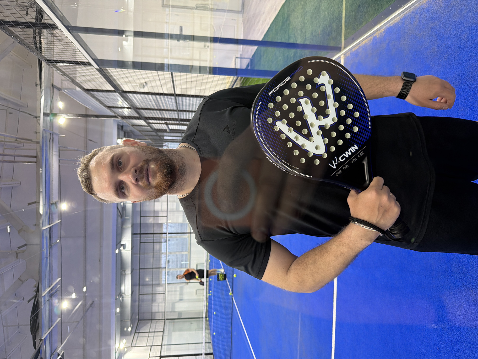 The image shows a Caucasian man holding a padel racket. The man has short brown hair, a beard, and is wearing a black t-shirt, a gold chain, a watch, and a bracelet. He is standing on a blue padel court with white lines. In the background, another person can be seen playing padel. Padel balls are scattered on the court near the net. The court is indoors with overhead lighting and a surrounding cage. The location is most likely a padel court in the city of Chaikovsky, Russia.