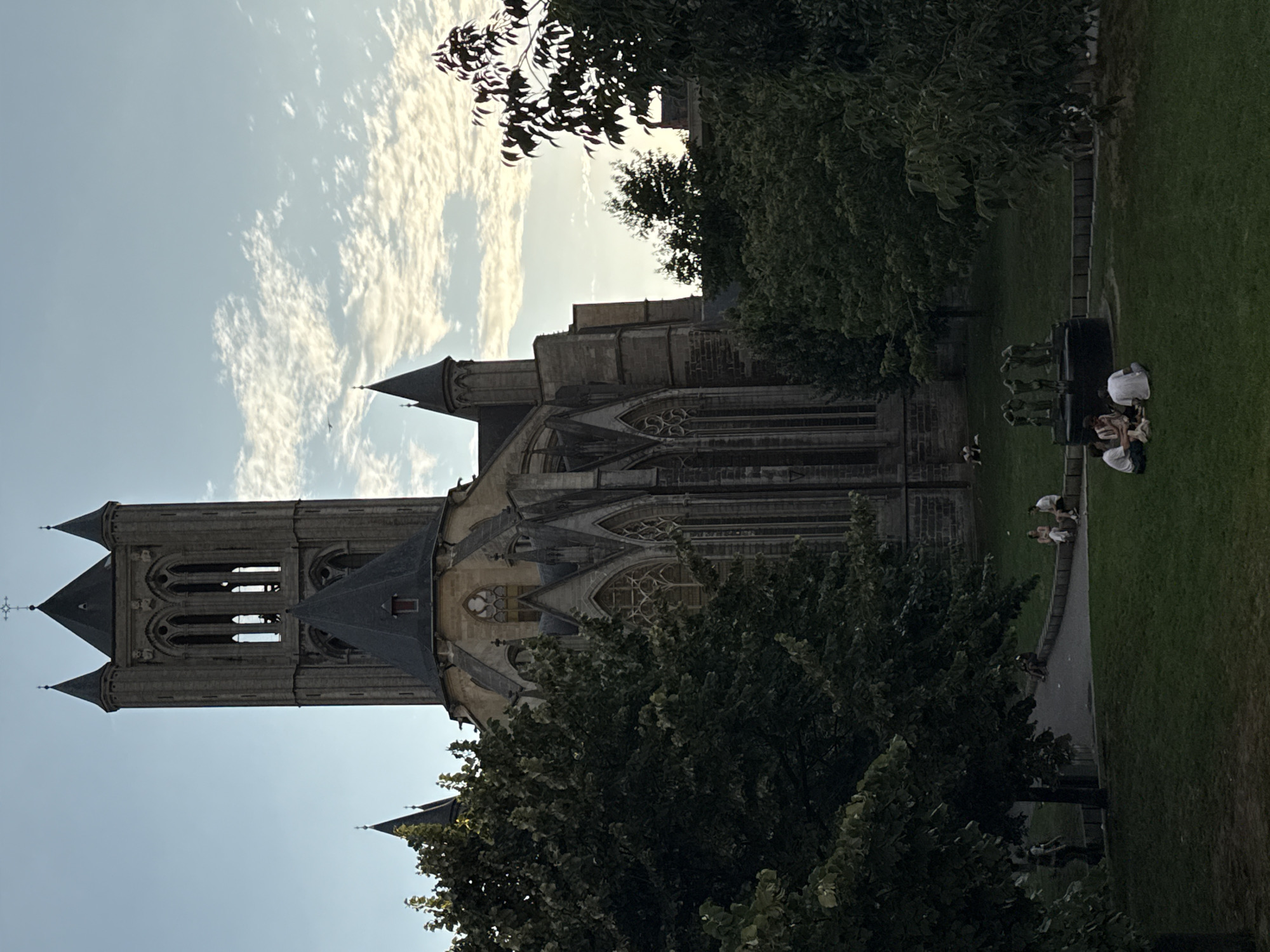 The photograph captures an outdoor scene at St. Bavo's Cathedral in Ghent, Belgium. The imposing Gothic architecture of the cathedral dominates the background, with its tall bell tower, spires, and intricate stonework clearly visible. Lush green trees frame the right side of the cathedral, their leaves creating a dappled shade. In the foreground, a grassy lawn is dotted with several groups of people. Some are seated, seemingly conversing or relaxing, while others are walking. A sculpture of several figures stands on a plinth in the middle ground, near a pathway. The sky above is a pale blue, with scattered clouds and a hint of sunset lighting.
