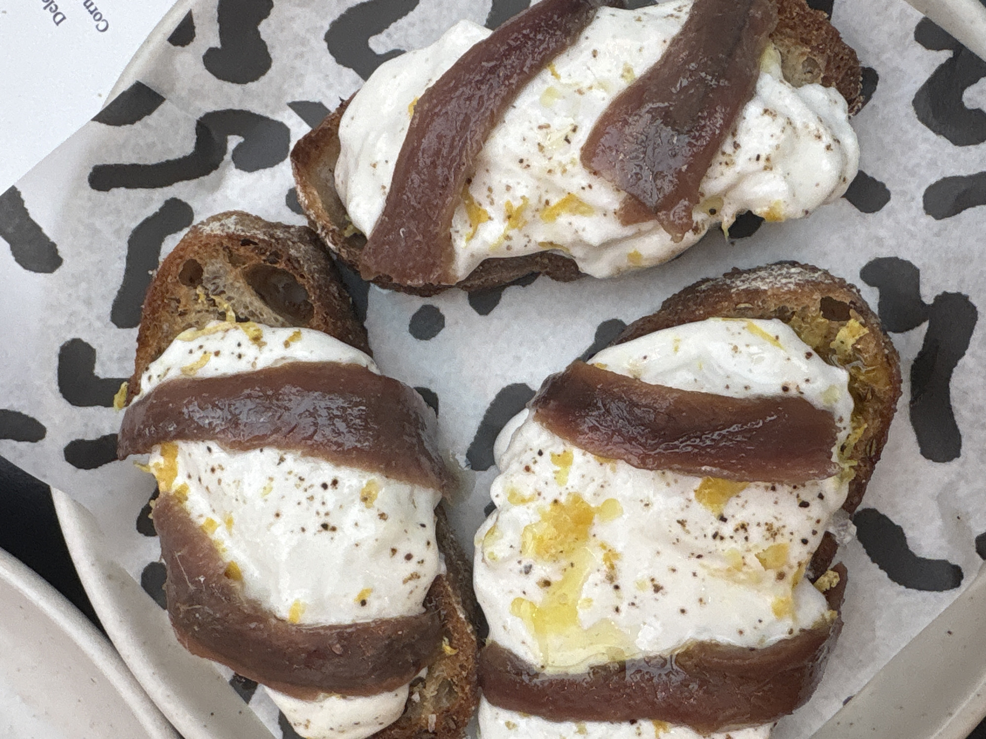 The image shows a plate of three appetizers, likely consisting of toasted bread topped with a creamy white spread, drizzled with oil, and decorated with dark brown anchovies. The plate is white with black, amoeba-like patterns. A portion of a white bowl or plate is visible on the left side. The scene appears to be in a restaurant. While the image doesn't explicitly show the location in London, United Kingdom, it may be a restaurant in that area.