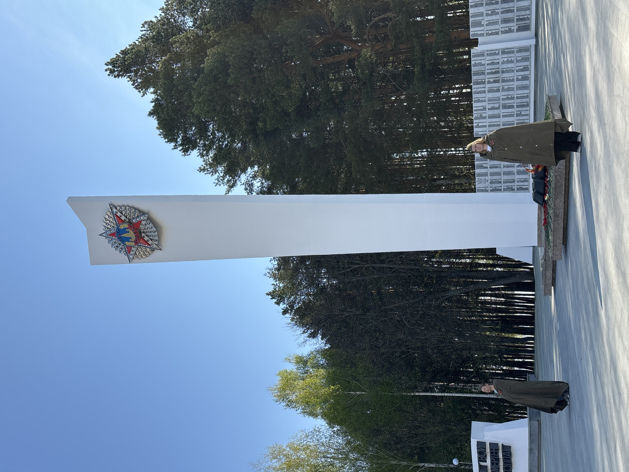 A photograph captures a view of the Memorial Complex to Soldiers Who Died in the Great Patriotic War, located in Chaikovsky, Russia. The monument is characterized by a tall, white, rectangular structure adorned with a red and silver Soviet star emblem. A woman stands near the base of the memorial, appearing to pay respects or place flowers. The background features dense trees and a clear, blue sky. White, block-like structures bearing inscriptions are also visible.