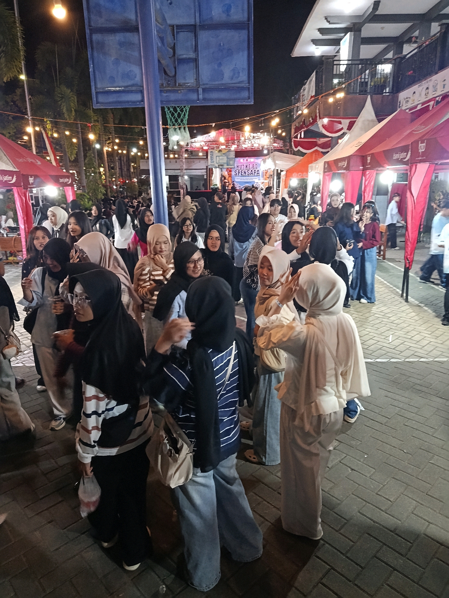 The image depicts an outdoor night scene in Pacitan, Indonesia, likely a public gathering or event. A crowd of people, primarily young adults, are visible, many wearing hijabs, indicating a predominantly Muslim population. They are gathered in an area with temporary stalls or tents, suggested by the red and white canopies. String lights are illuminated, creating a festive atmosphere. A basketball hoop and backboard are partially visible in the background. The ground is paved with interlocking bricks. Several individuals appear to be interacting, talking, or taking photos. The overall impression is of a lively community event in a city setting at night.