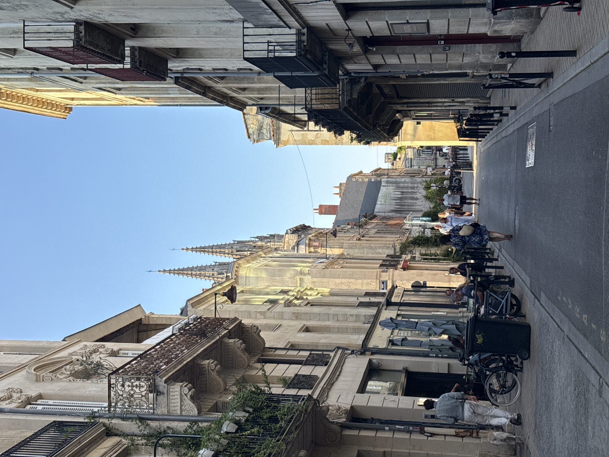 A medium shot shows a street in Bordeaux, France, lined with tall, historic buildings. The buildings feature ornate balconies, stone facades, and shuttered windows. In the distance, the spires of a cathedral are visible against a clear blue sky. Several people are walking or sitting at outdoor tables in front of cafes. Two bicycles are parked near a large trash receptacle on the sidewalk. The street is paved with asphalt, and a cobblestone sidewalk runs alongside the buildings. The lighting suggests it is late afternoon or early evening, with the sun casting long shadows.