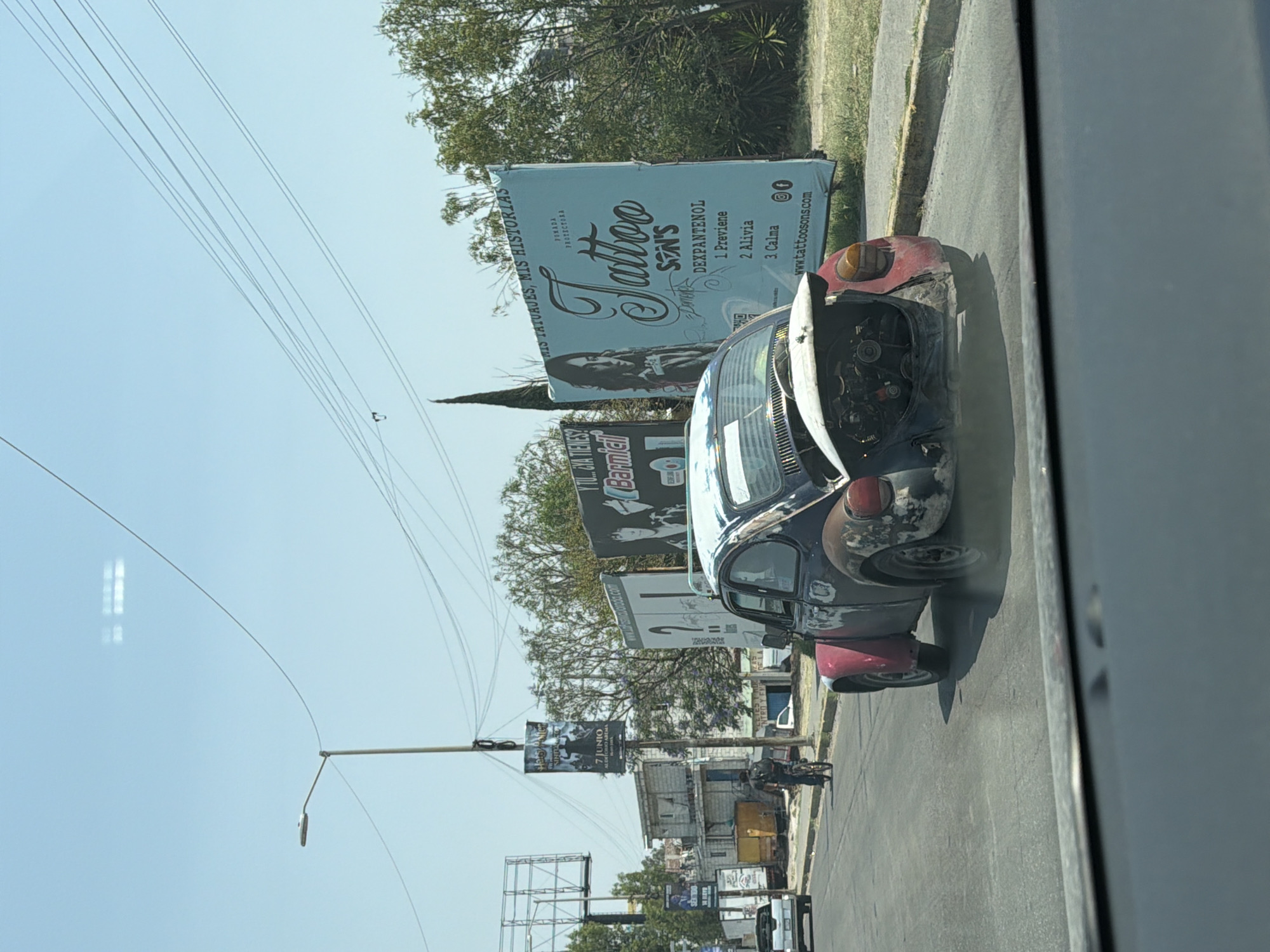 The image, taken from inside a vehicle, shows a partially-disassembled black and red Volkswagen Beetle with its hood open, parked on the side of a road in San Bernardino Tlaxcalancingo, Mexico. Various billboards are visible in the background, one advertising "Tattoo Sons" with the phrase "Mis Tatuajes, Mis Historias," indicating a tattoo parlor. The environment includes utility poles and wires overhead, trees, and other buildings along the street. Another billboard advertises a June 7th event at Arena.