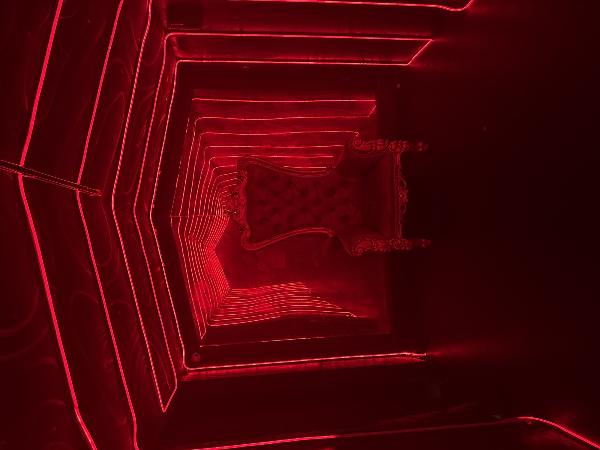 This image captures an interior space, likely a corridor or a specially designed room, characterized by pervasive red lighting. The primary subject is an ornate, upholstered armchair, resembling a throne, positioned at the end of what appears to be a tunnel or a recessed alcove. The chair features intricate carvings on its wooden frame and is upholstered in a tufted red fabric.
The setting is defined by numerous parallel lines of red neon or LED strip lights that run along the walls, ceiling, and floor, creating a strong sense of perspective and depth. These lights illuminate the path leading towards the grand chair, intensifying the red hue throughout the scene. The surfaces, possibly reflective walls or a polished floor, amplify the light, contributing to the dramatic and immersive atmosphere.
No human activity is visible, making the scene static and drawing full attention to the interplay of light and form. The strong red lighting evokes a sense of intensity, luxury, or a vibrant nightlife environment. The opulent chair suggests a focal point, an invitation, or a statement piece within this unique space. There is no visible text. Given the context, this striking installation is located in North Kuta, Indonesia.