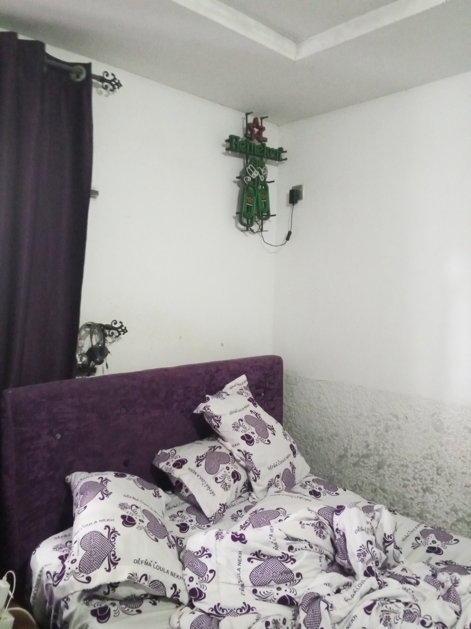 The image depicts a bedroom interior, likely in Abuja, Nigeria. The scene is dominated by a large, plush purple headboard and matching bedding. The bed is covered with white duvet and pillows, adorned with a repeating pattern of purple hearts and floral motifs. Some of the pillows have text written on them, but it is illegible.
On the white wall behind the bed, there is a neon sign shaped like a star and the word "Heineken" in green lettering, suggesting a possible bar or man cave theme. Next to the sign, a black electrical outlet is visible. To the left of the neon sign, a decorative metal piece with intricate scrollwork hangs on the wall, and a dark purple curtain is drawn to the side, partially obscuring the corner of the room.
The wall texture changes from smooth white paint to a rougher, greyish finish towards the right side of the image. The ceiling is white and features recessed lighting. The overall impression is a lived-in, personal space, likely a bedroom in a residential setting in Abuja. There are no people visible in the image.