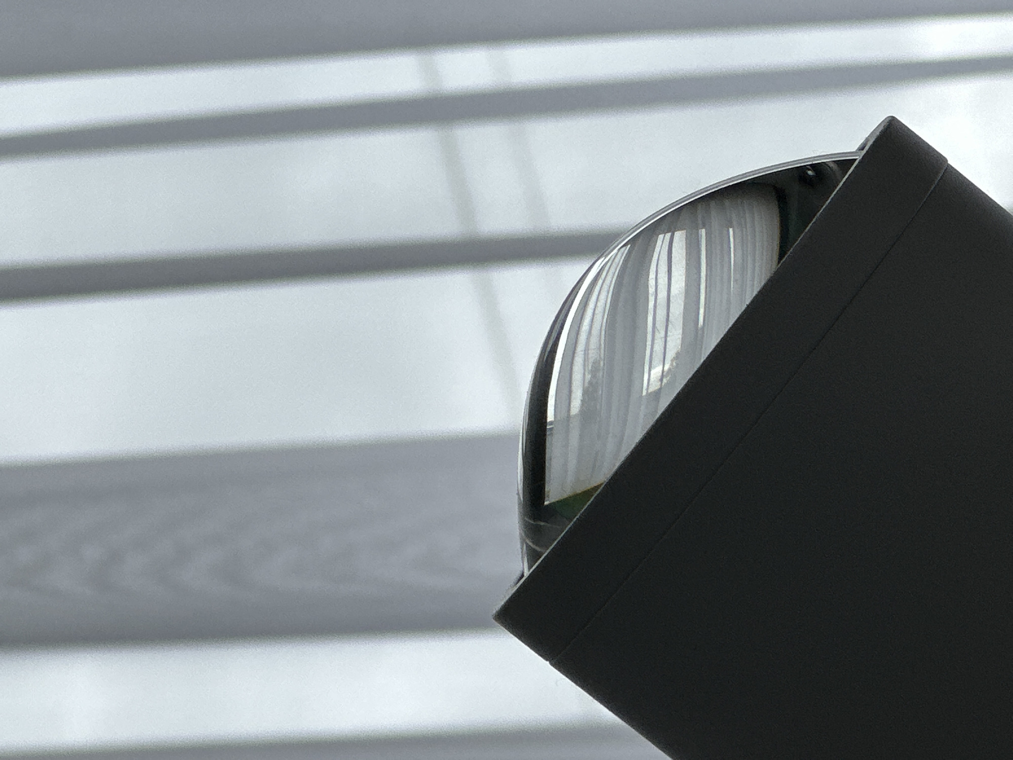 The image shows a close-up of a dome-shaped object with a black rectangular casing partially obscuring it. The dome appears to be transparent or reflective, with vertical lines visible within. The background consists of horizontal, light-colored stripes, potentially blinds or slats. The location is Unknown, Unknown.
