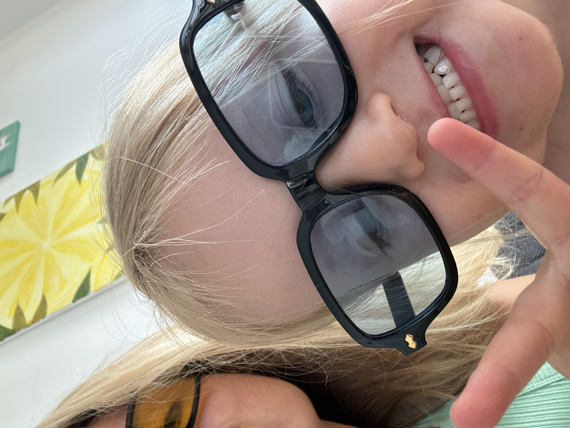 This is a close-up, over-the-shoulder selfie taken outdoors in the city of València, Spain. In the foreground, a young girl with blonde hair is wearing large, black-framed glasses. She is smiling and making a peace sign with her left hand. To the left of the girl, another person with blonde hair and yellow-tinted sunglasses is partially visible. Behind them, a painting with yellow and green elements is hanging on a white wall. The image appears to be taken during daylight.