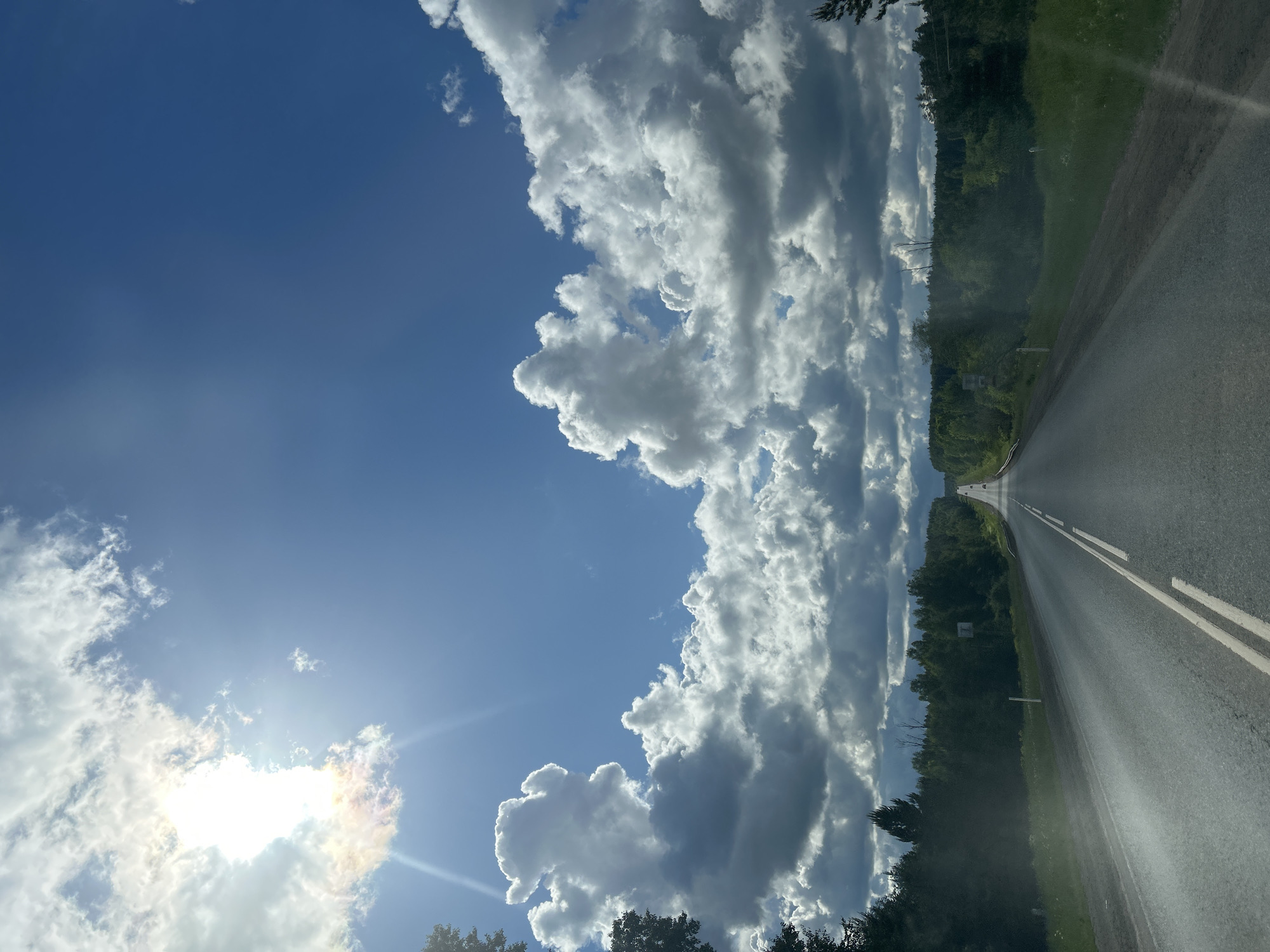 The original image, which is rotated 90 degrees counter-clockwise, depicts a scenic view from a road on a sunny, partly cloudy day. When viewed correctly, a long, straight asphalt road extends from the bottom center into the far distance, marked by a prominent double white line in the middle and a single white line on its right edge. The road surface appears somewhat damp, reflecting light.
Dense green forests line both sides of the road, forming a verdant corridor under the sky. The trees are tall and healthy, contributing to the natural, expansive setting.
The sky is a bright blue, particularly on the left side of the frame, but it is dramatically filled with large, puffy cumulus clouds of white and grey on the right side, extending vertically from the horizon upwards. The sun is clearly visible in the upper left portion of the sky, partially hidden behind a cloud, emitting bright rays and lens flare.
In the far distance along the road, faint outlines of other vehicles can be discerned, along with a small white marker pole on the right shoulder and an indistinct road sign further away. The scene captures a moment during a drive, likely in a rural or intercity area near Chaikovsky, Russia. The time of day appears to be midday due to the bright sunlight, and the weather is characterized by a clear sky interspersed with striking cloud formations. No legible text is visible in the image.