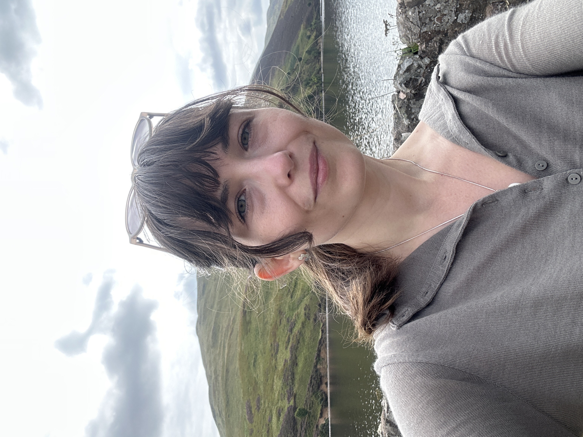 A young woman with brown hair and bangs, wearing a grey sweater and a silver necklace, takes a selfie outdoors. She is positioned in the foreground, with a body of water and rolling green hills in the background. The sky above is cloudy, with a hint of sunlight breaking through. The location appears to be a natural landscape, possibly a reservoir or lake, in the vicinity of Penicuik, United Kingdom.