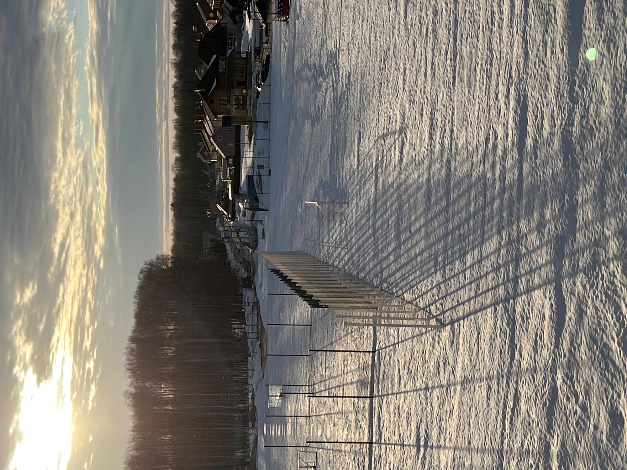 The image presents an outdoor winter scene in Noviy, Russia, observed under low-angle sunlight, indicating either sunrise or sunset. The sky displays a varied cloud cover, with bright illumination in the upper left quadrant originating from the partially obscured sun, transitioning to darker cloud formations towards the right.
The immediate foreground and midground consist of a vast expanse of snow-covered terrain, exhibiting a textured, undulating surface. Within this area, two basketball hoops with backboards and rims are visible on the left side, accompanied by numerous dark vertical poles arranged in a grid. A light-colored fence, constructed from vertical slats or posts, diagonally traverses the field from the lower left towards the mid-right, casting prominent, elongated parallel shadows across the snow. A single, light-colored bench or seating object rests on the snow to the right of the basketball hoops, also casting a shadow. No human or animal figures are present. A small, bright green circular light artifact is present in the lower right corner of the frame.
In the background, a dense line of bare deciduous trees forms a dark, linear silhouette. Beyond the tree line, a residential development is visible, featuring multiple detached houses. These structures are primarily two-story, with varying architectural styles and predominantly reddish-brown or dark-colored pitched roofs. Scattered vehicles, some resembling boats on trailers, or other recreational equipment are discernible within the residential property boundaries. The overall scene is static, devoid of active human or animal interaction.