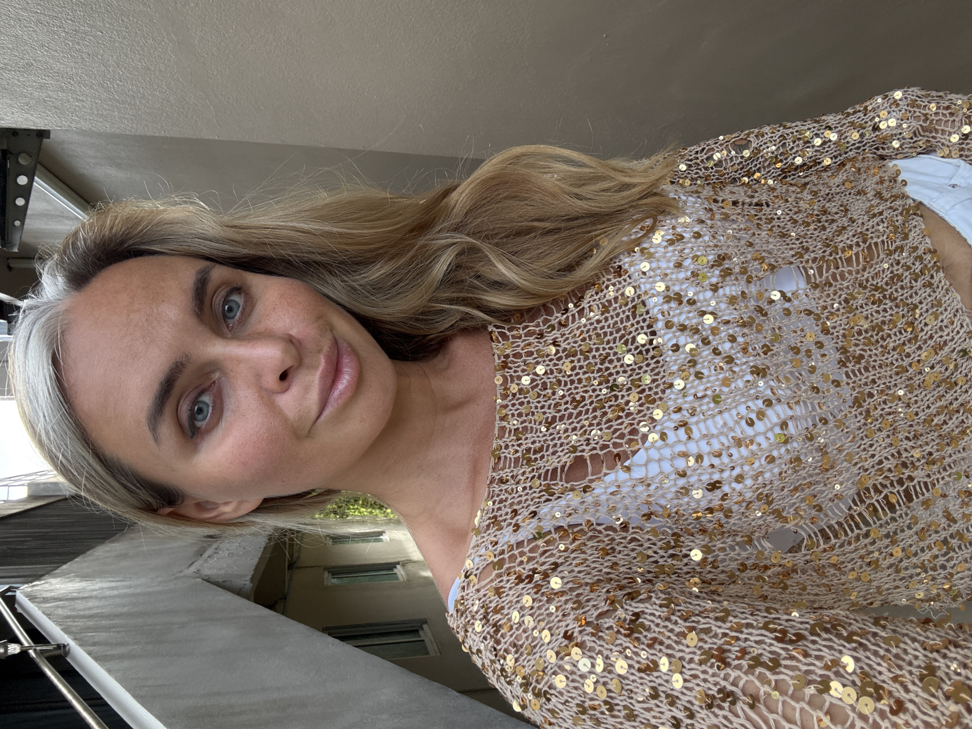 A medium shot of a woman standing outdoors, facing the camera. She has blonde hair, blue eyes, and is wearing a sparkly gold top over a white garment. The background shows a concrete structure with stairs and some greenery. The wall behind her is a neutral color. The scene appears to be in an urban or residential area.