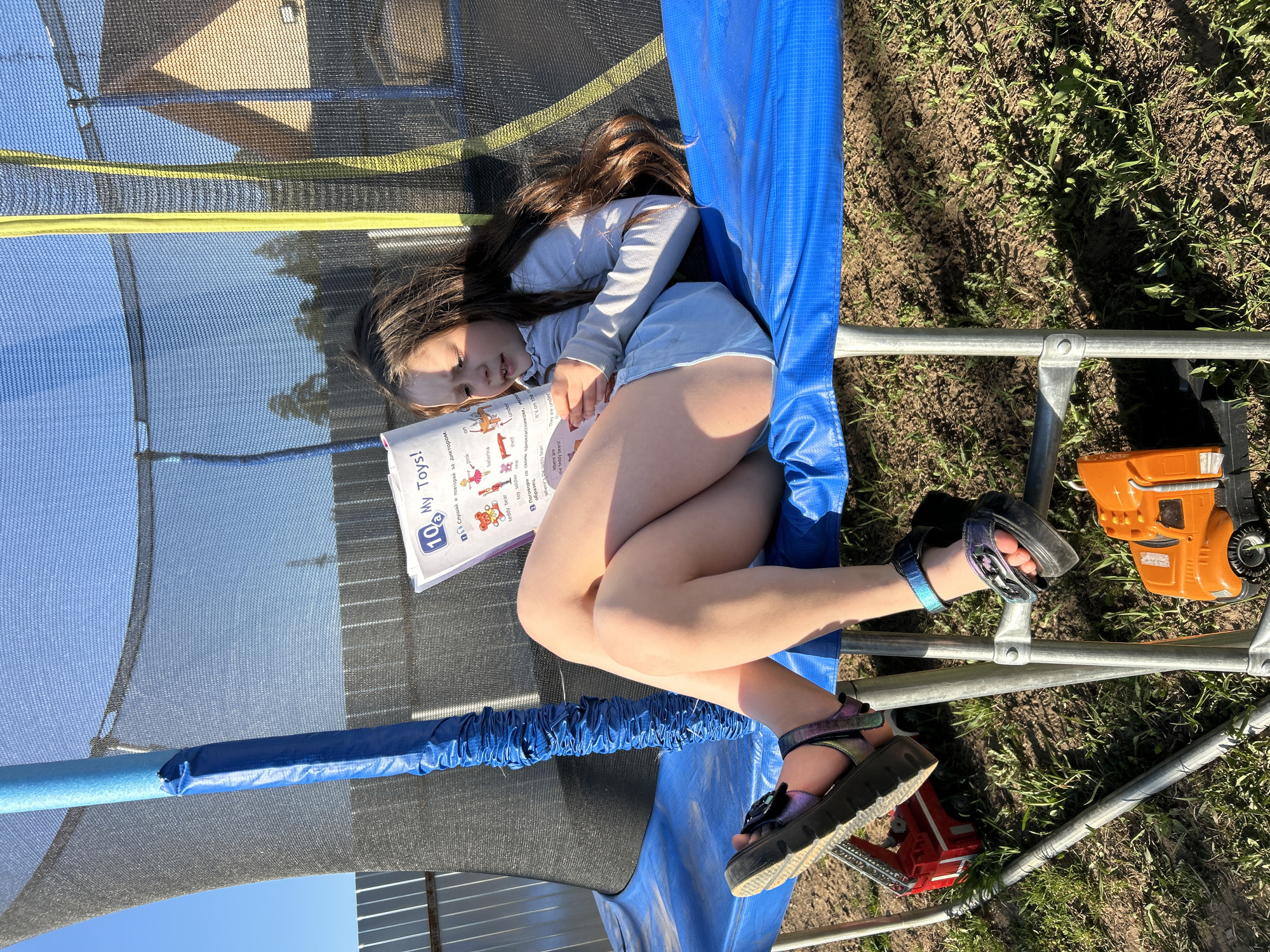 A girl, approximately 8-10 years old, is lying on a trampoline. She has long brown hair and is wearing a light-colored long-sleeved shirt and light blue shorts. She is holding a book titled "10 My Toys!" and appears to be reading. Her feet are adorned with sandals. The trampoline has a blue padded edge and safety netting. On the ground next to the trampoline are a red toy fire engine and an orange toy truck. The background shows grass, partial trees, and a patch of blue sky. The OCR text within the image indicates that the book is in Russian. The location and city of Chaikovsky, Russia, are not discernible from the image.