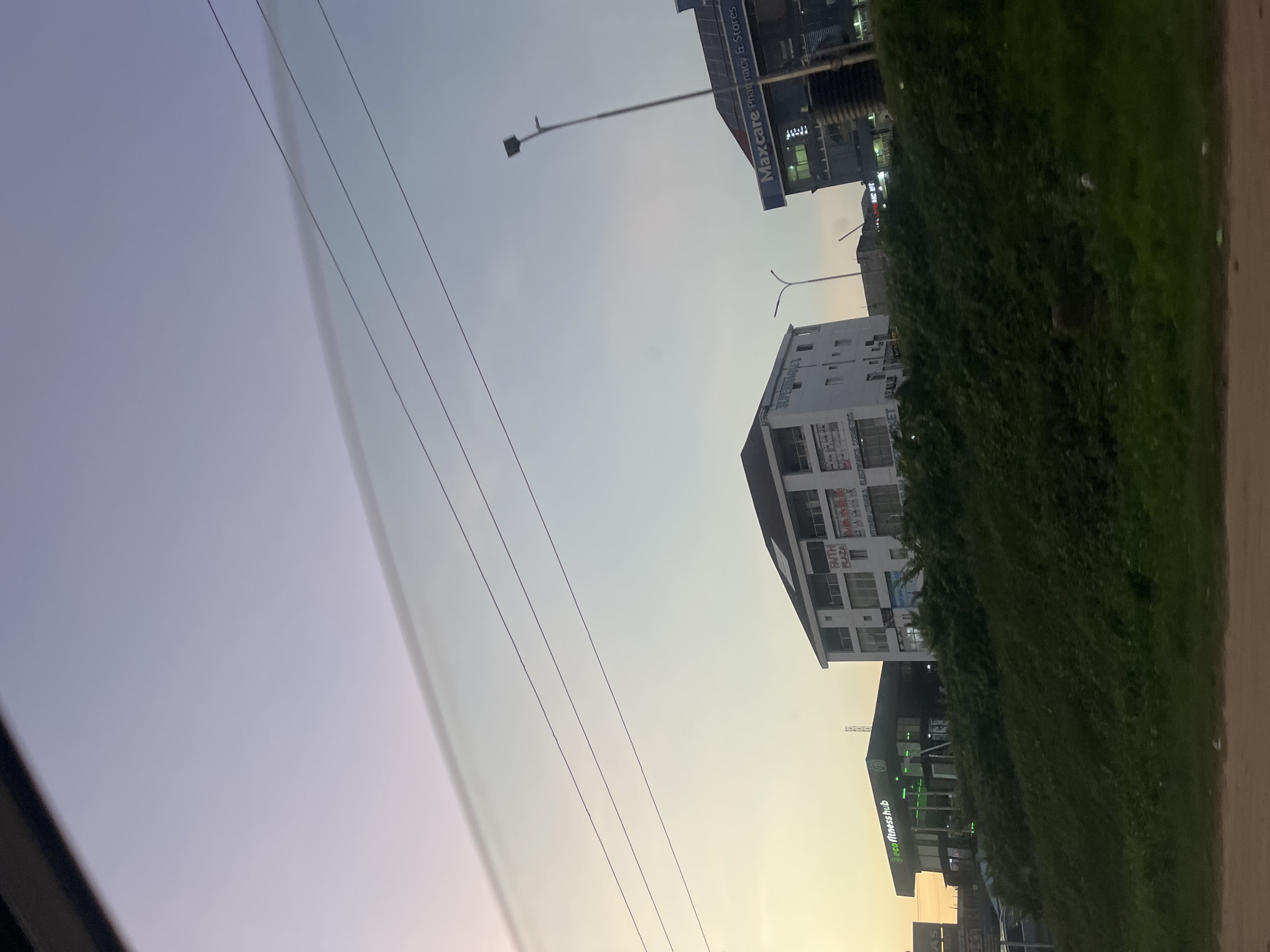 This image captures a street scene in Galadimawa Village, Nigeria, depicting several commercial buildings against a twilight sky. The sky transitions from a soft yellow-orange at the lower left horizon to light purple and pale blue at the top. Multiple overhead power lines traverse the upper middle section of the frame. A distinct, elongated cloud formation runs horizontally across the sky, parallel to and above some power lines.
In the mid-ground, from left to right, are three prominent buildings. On the far left, partially obscured by a dark foreground, is a modern structure identified as "eco fitness hub" with green illuminated signage. Adjacent to it on the right stands a multi-story white building with multiple balconies. A sign near its roof reads "FAITH PLAZA." Other signs on its lower levels include "SUPERMARKET," "ELECTRONICS & COSMETICS," and partially visible "KITCHEN UTENSILS & ELECTR...". To the right of this building is a dark blue commercial structure with a prominent white and blue sign displaying "Maxcare Pharmacy & Stores."
The foreground consists of a dark green, overgrown grassy slope that rises towards the buildings, suggesting an elevated median or roadside embankment. A dirt road or shoulder is visible at the very bottom of the frame. Scattered streetlights and utility poles are visible throughout the mid-ground, with one tall streetlight positioned prominently to the right of the central white building. No individuals are discernible within the frame.