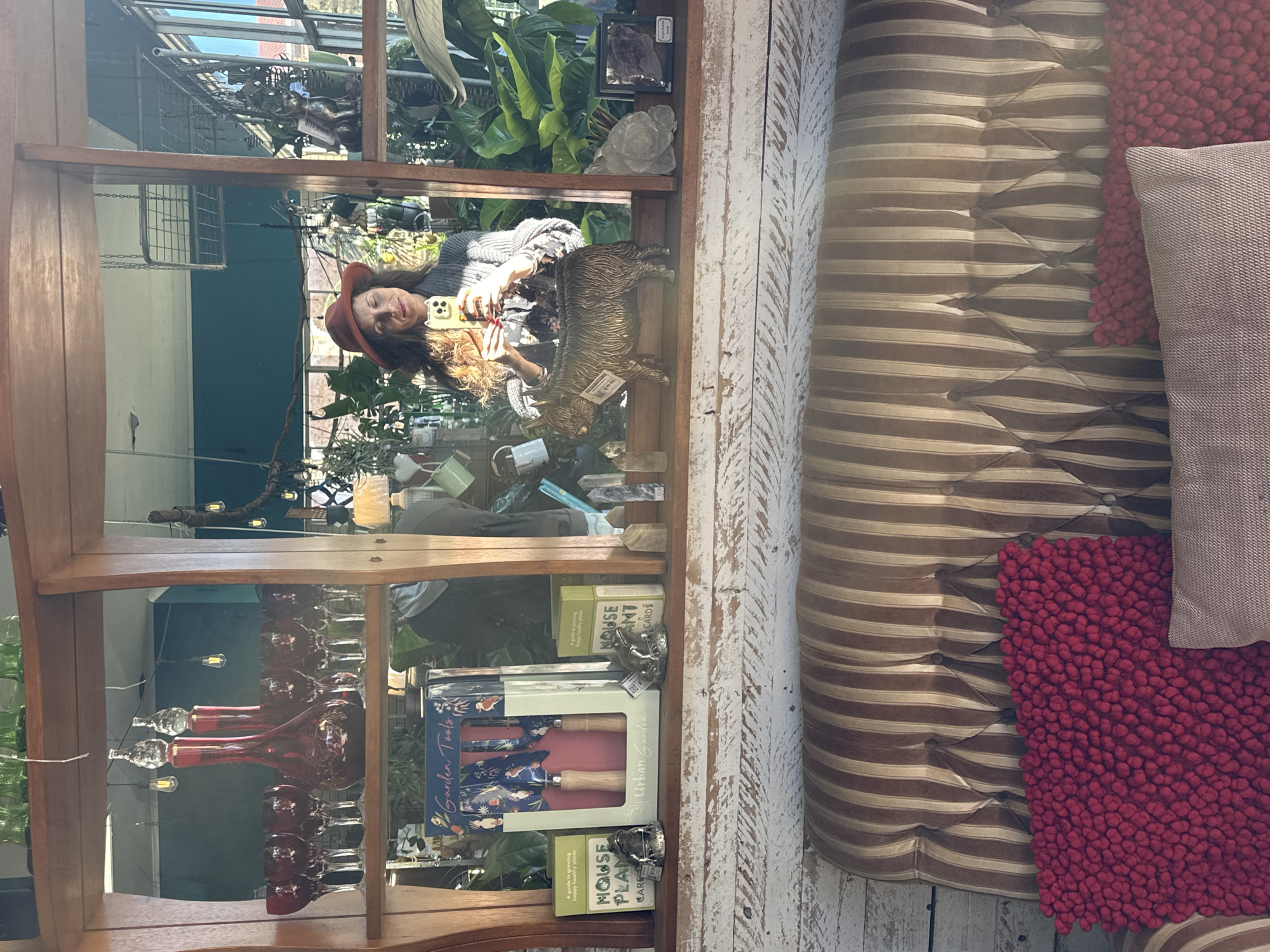 The image shows a woman taking a selfie in a mirror, reflecting a room filled with plants and decorative items. She is wearing a red hat and a grey knitted cardigan over a floral top. In the foreground, a tufted brown and beige striped sofa is visible with two pillows on it, one red and textured, the other a light pink. The background in the mirror reveals a shelf with red glass decanters and wine glasses, alongside boxes labeled "Garden Tools" and "House Plant Cards". A bronze figurine of a highland cow is also on the shelf. The environment appears to be a retail space or a home decorated with plants and vintage items. The location is specified as Bellingen, Australia, likely a shop or an apartment in the town.
