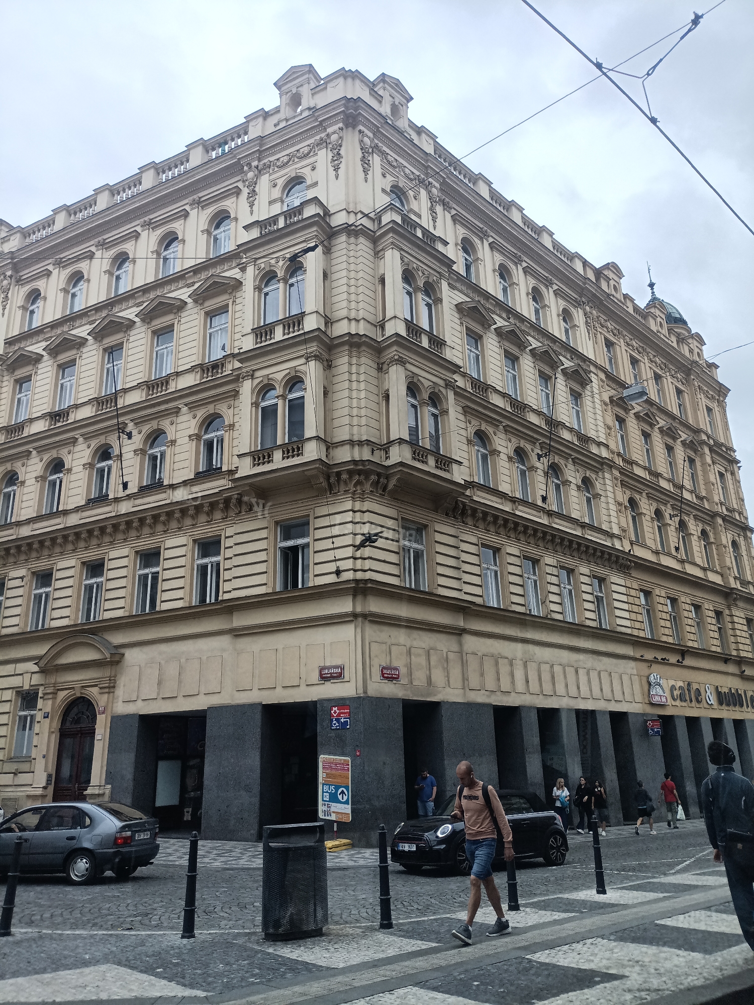 The image depicts a street scene in Prague, Czechia, with a focus on the corner of an ornate, multi-story building. The building's facade is a light cream color, featuring numerous arched and rectangular windows with white frames, separated by decorative columns and horizontal bands. The architecture suggests a style common in Central European cities.
In the foreground, a cobblestone street is visible, with a crosswalk featuring a black and white pattern. Several bollards are positioned along the sidewalk edge.
On the street, a dark-colored sedan is parked on the left side. A black convertible is also present, with a person walking in front of it. A man in a light-colored long-sleeved shirt and denim shorts is walking across the crosswalk. Another person, wearing a dark jacket, is walking away from the camera on the right side of the street. Other pedestrians are visible further down the street, near the building.
The ground floor of the building has a darker facade with large, recessed openings, one of which appears to be an entrance. A sign above one of these openings reads "cafe & bubble". Street signs are visible on the building, including one with "JUGOSLAVSKA" and another with a metro symbol and accessibility icon. A blue and white sign with bus information is also present. A black trash receptacle stands near the sidewalk. Overhead, electrical wires are strung across the sky, indicating the presence of a tram or trolley system. The sky is overcast.