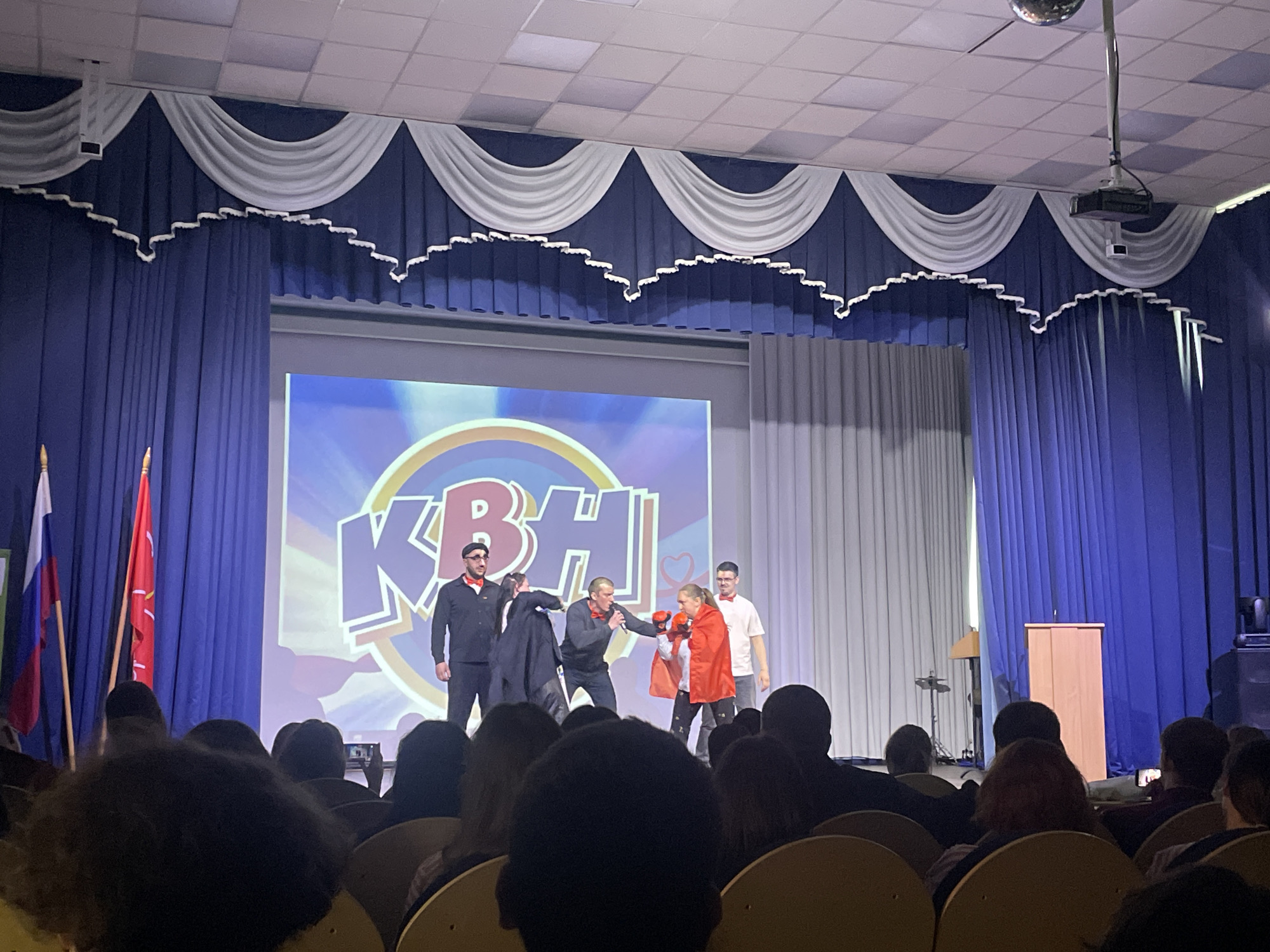 A medium shot shows a performance on a stage in what appears to be a school or community hall in Novoe Devyatkino, Russia. The stage has blue curtains with white draping at the top. A large screen behind the performers displays the "KBH" logo.
Five performers are on stage. From left to right: one man in a dark uniform-like suit with a red bow tie and beret, a man in a gray sweater pushing another man in a black coat, a woman in an orange cape and boxing gloves, and a man in a white shirt and red bow tie. The woman appears to be preparing to box.
An audience is seated in rows of chairs facing the stage. Two flags, one of which is the Russian flag, are seen on the left side of the stage. A lectern is on the right side of the stage. A projector and a disco ball are visible on the ceiling.