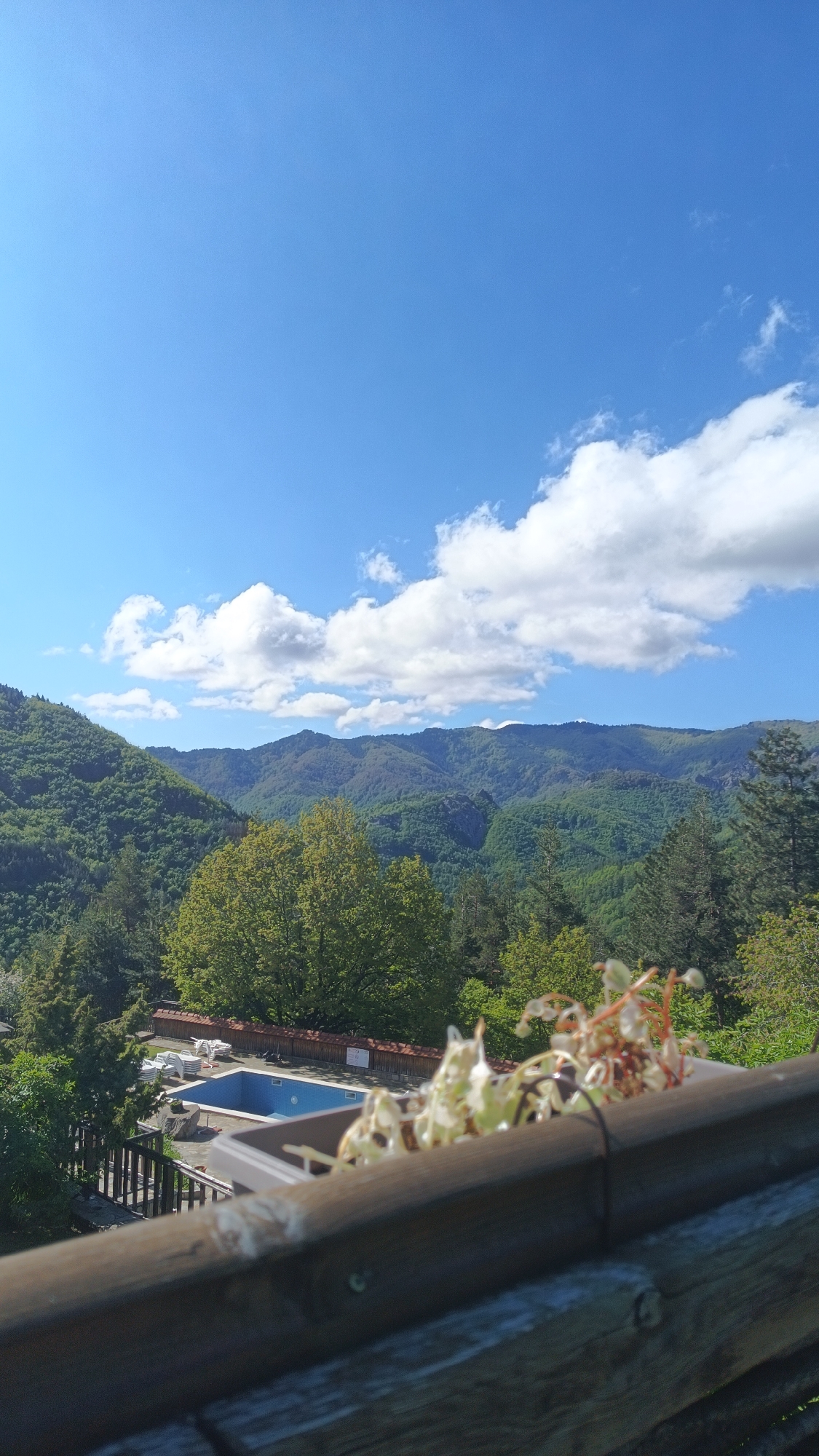 The image shows a view from a balcony in Vrata, Bulgaria, overlooking a landscape featuring a swimming pool, lush green mountains, and a partly cloudy sky. In the foreground is a wooden railing with a planter containing dried flowers. The swimming pool area includes lounge chairs. Beyond the pool, the densely forested mountains are visible under the blue sky dotted with white clouds.