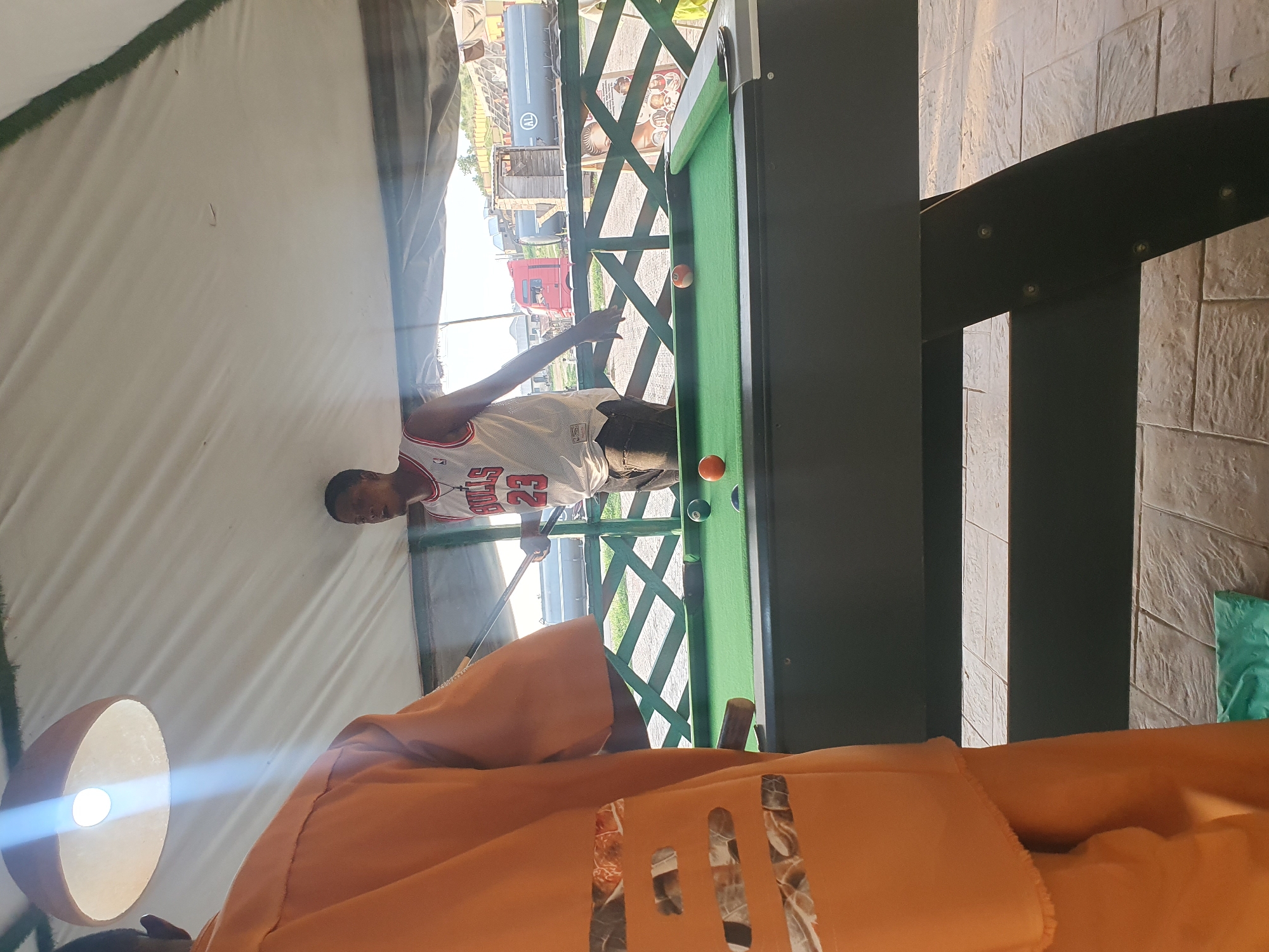 In Lokoja, Nigeria, two individuals are engaged in a game of billiards outdoors under a tented structure. One person, seen from the back and wearing an orange garment with patterned cutouts, stands to the left. The other individual, facing the billiard table, wears a white Michael Jordan Chicago Bulls jersey with the number 23 and dark trousers. The billiard table is green, with several balls visible, including one orange ball. A cue stick is held by the person in the Bulls jersey. In the background, a green lattice fence separates the play area from a view of large tanker trucks and buildings. The ground is paved with textured square tiles. Overhead, a light fixture hangs from the tent.