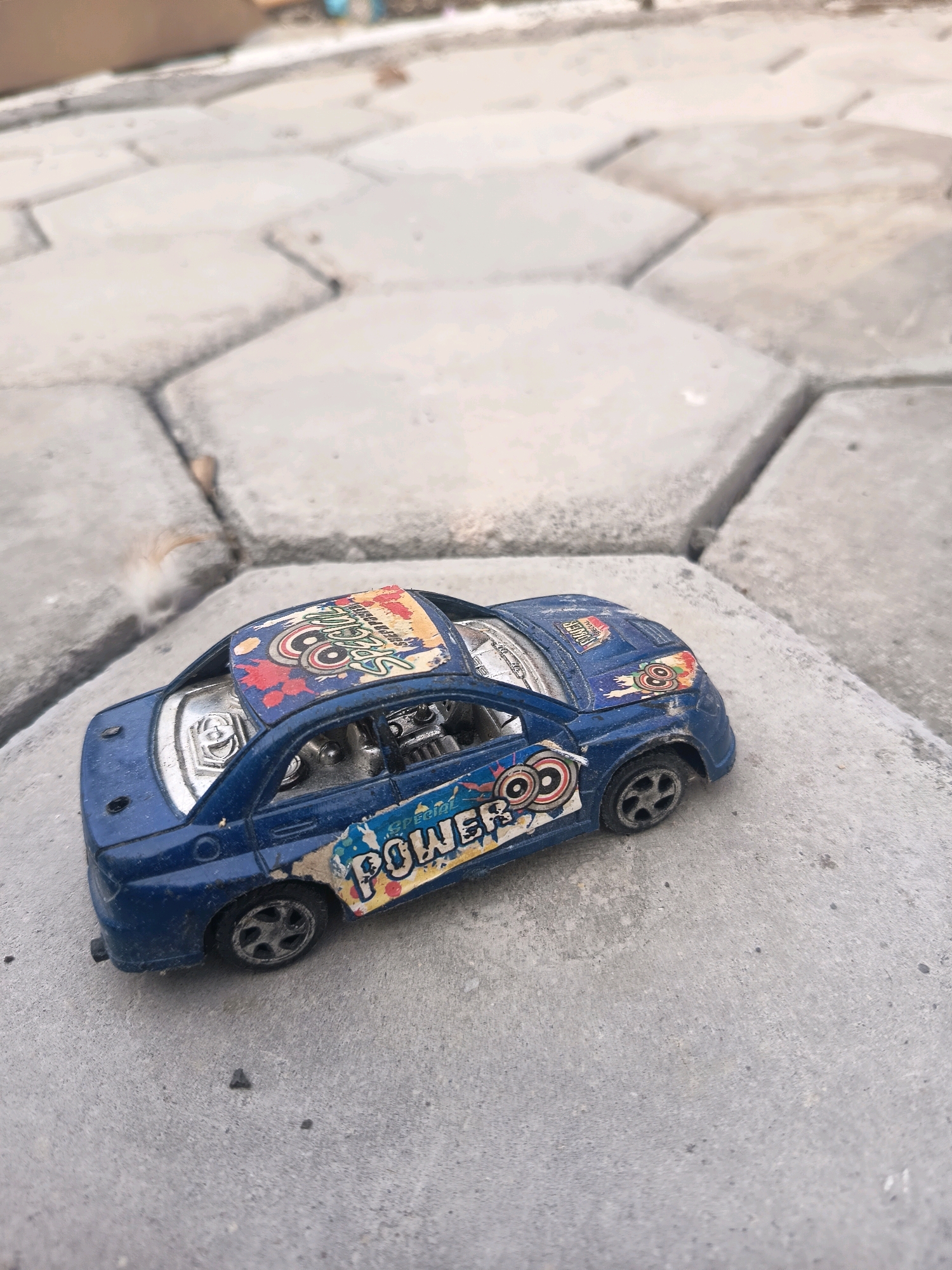 A blue toy car with colorful decals sits on a concrete surface. The car has the word "POWER" written on its side. The concrete surface is composed of hexagonal pavers. In the background, the scene is blurred and suggests an outdoor environment. The image appears to have been taken at ground level. There are no people or animals visible in the image. The lighting is natural and appears to be daylight.