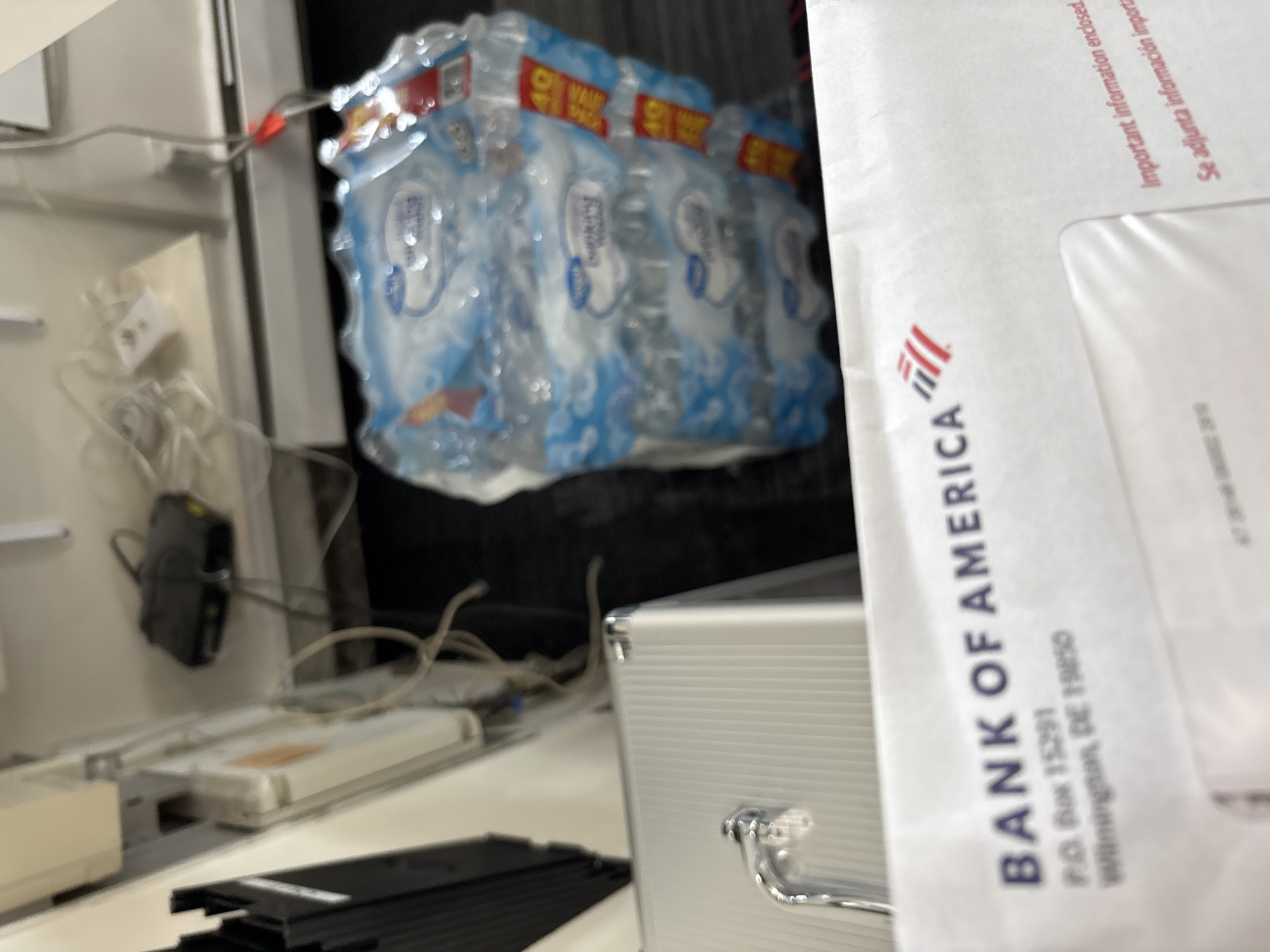 An indoor, medium-shot photograph displays a collection of objects, primarily focused on an envelope and packaged bottled water, against a backdrop of electronic devices and cables. In the right foreground, a white envelope from "BANK OF AMERICA" is prominently visible, oriented vertically. Visible text on the envelope includes "BANK OF AMERICA", "P.O. Box 15291", and "Wilmington, DE 19850". Further up, "Important information enclosed" and "Se adjunta información import." (Spanish for "Important information attached") are also legible.
Behind the envelope and slightly to the left, a silver-colored metallic case with a handle is partially visible. In the upper mid-ground, a multi-pack of bottled water, likely a 24-pack, is stacked. The bottles have blue labels, and red and yellow tags indicating "40" and "VALUE" are attached to the packaging. The bottles appear to be a store brand, possibly "Everyday Essentials" or similar, though text on the bottles is blurry.
To the left of the scene, various electronic components and tangled white and black cables are present, including a black device hanging from what appears to be a shelf or a wall-mounted hook, and a white power adapter. A long, white rectangular device with some orange labeling is also visible on a surface, likely a desk or counter. The background behind the bottled water is dark, suggesting a cabinet or a shadowed area. The overall impression is one of a cluttered utility space or a back office in Hampton, United States of America. No people are visible, and no specific activity is taking place. The lighting suggests indoor artificial illumination. The image has a shallow depth of field, with areas of significant blur.