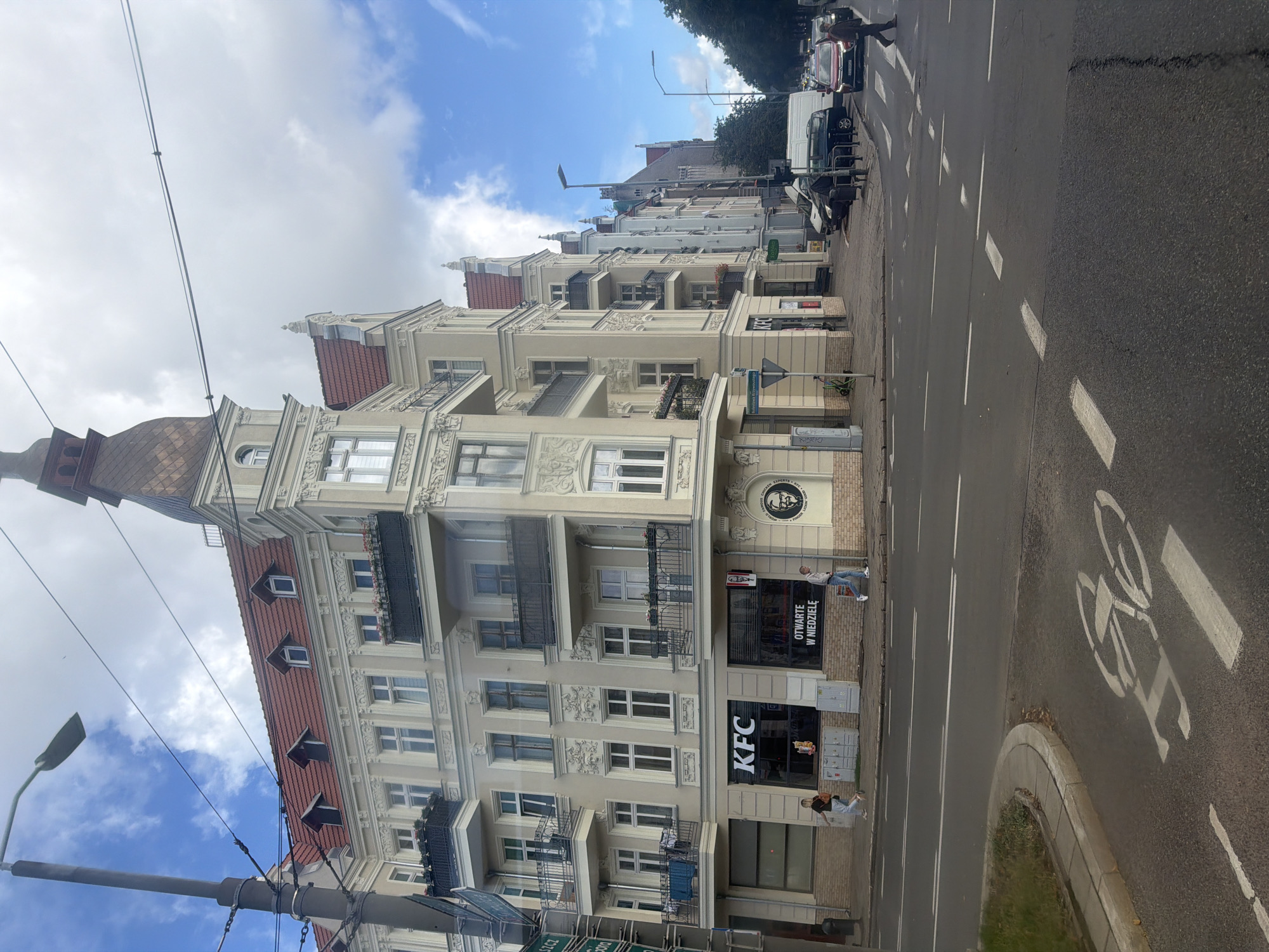 The image depicts a street scene in Szczecin, Poland, specifically at the intersection of Monte Cassino Street and Bohaterów Getta Street. A multi-story building with a distinctive tower and red-tiled roof dominates the left side of the frame. The building features ornate architectural details, balconies with black railings, and several windows. On the ground floor, a KFC restaurant is visible with its prominent signage. Two individuals are walking on the sidewalk in front of the KFC. The street in the foreground is asphalt with lane markings and a marked bicycle lane. Several cars are parked and driving on the street, including a white van and a red car. The sky above is partly cloudy with patches of blue sky. In the background, other buildings and trees are visible.