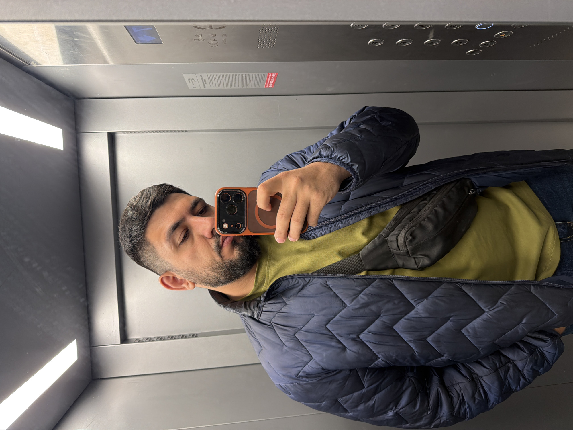 A man with short, graying hair and a beard is taking a selfie in an elevator. He is wearing a dark blue puffer jacket, a greenish-yellow shirt, and a black fanny pack. The elevator has a metal interior with a rectangular light panel on the left and a control panel on the upper portion. The elevator is located in Krasnoyarsk, Russia, as indicated by text on the control panel.