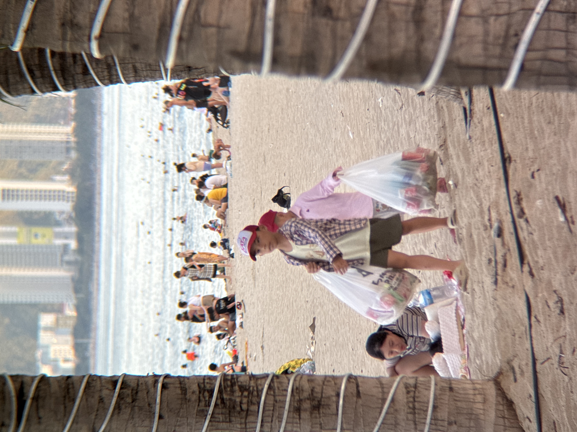 The image captures a crowded beach scene in Nha Trang, Vietnam, likely during daylight hours given the bright conditions. In the foreground, partially obscured by palm tree trunks, two children are visible on the sand. The child on the left, wearing a plaid shirt and shorts, carries a large clear plastic bag filled with items. The child on the right, wearing a pink long-sleeved shirt and shorts, also holds a similar bag. A woman is crouched on the sand to the left, appearing to be interacting with something on the ground, with a water bottle and a patterned mat nearby.
In the midground, the beach is populated with numerous people, some wading in the shallow water, others sitting or standing on the sand. The ocean stretches out towards a backdrop of tall, modern buildings, indicating the urban setting of Nha Trang. The water is a light blue, and the sand is a light brown, typical of a coastal environment. The overall atmosphere suggests a lively and busy beach day.