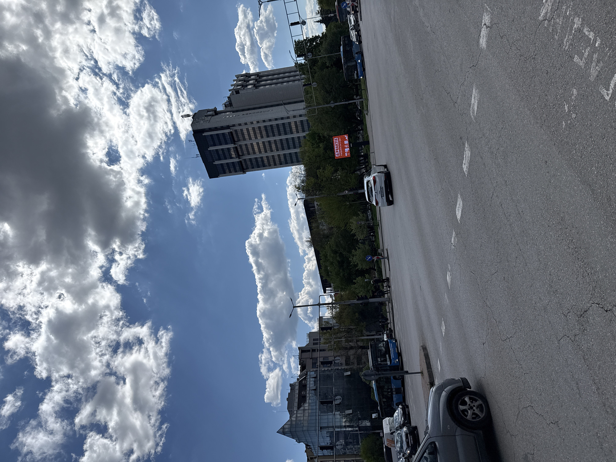 A bright, sunny street scene in a city, likely Plovdiv, Bulgaria, depicts daily urban activity under a vibrant blue sky with scattered white cumulus clouds.
The image, oriented vertically, shows a wide asphalt road with white painted markings and visible cracks extending into the distance. In the immediate foreground, a dark grey SUV is partially visible, parked or moving along the curb. Further down the road, a white SUV is driving, and a blue bus is seen alongside other parked vehicles.
To the left of the road stands a tall, imposing building with multiple stories, featuring a facade of windows and light-colored concrete panels. Cyrillic text, possibly "МИНИСТЕРСТВО," is visible horizontally across its upper section. Trees line the street in front of this building, providing shade. A red billboard with white Cyrillic text "ПРАКТИС" (PRACTIS) and "Направи си сам" (Do it yourself) is prominently displayed near the street in front of the tall building. Further down on the right side, a modern building with reflective glass is partially visible.
The clear blue sky, abundant white clouds, and visible shadows indicate bright daylight, likely mid-morning or early afternoon. The scene captures a typical day in a bustling city environment, with no specific event taking place beyond routine traffic and urban presence.