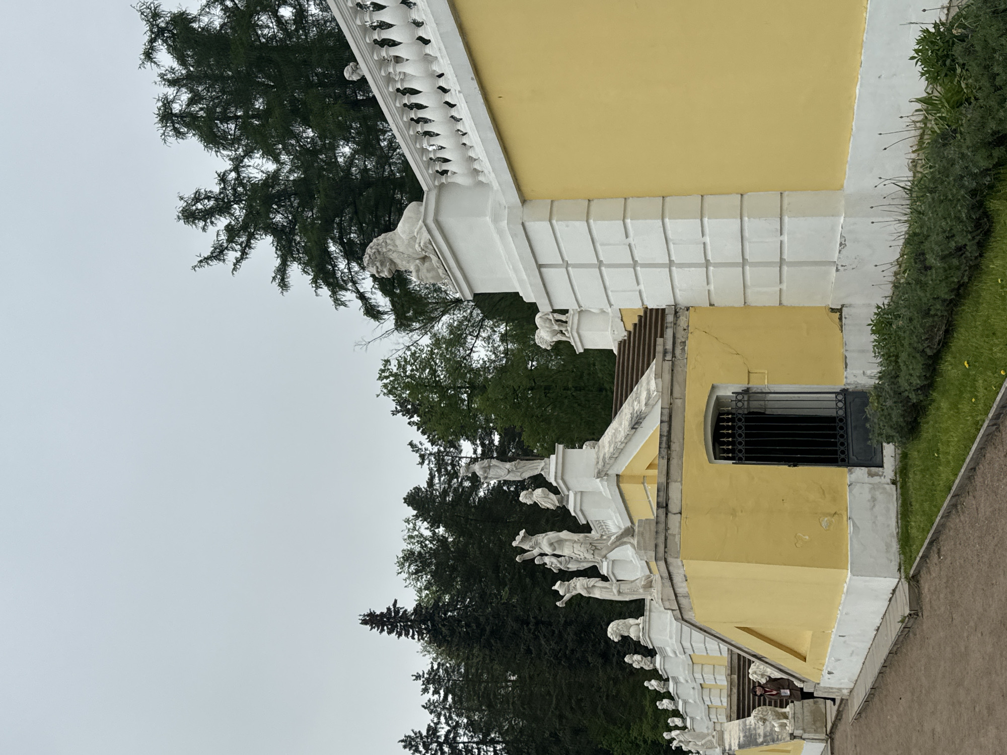 The image depicts a section of the palace in Arkhangel'skoe, Russia. The building is painted yellow with white architectural details, including a balustrade featuring sculpted figures. A window with a dark metal grate is visible. Trees and other greenery are present in the background. The sky is overcast.