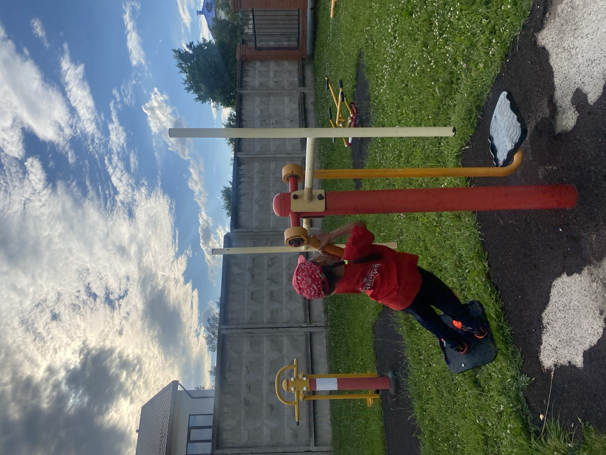 In Chaikovsky, Russia, a young girl is exercising outdoors on a bright, cloudy day. She is wearing a red hoodie and a red bandana, and is using a piece of outdoor gym equipment. The equipment appears to be a leg press machine. The girl is standing on a black platform, holding onto red handles. The scene is set in a park or playground, with green grass and a concrete fence in the background. The sky is filled with dramatic clouds, suggesting a dynamic weather system. The overall mood seems to be one of active play and enjoyment of the outdoors. The red hoodie has text on it, but it is not legible.