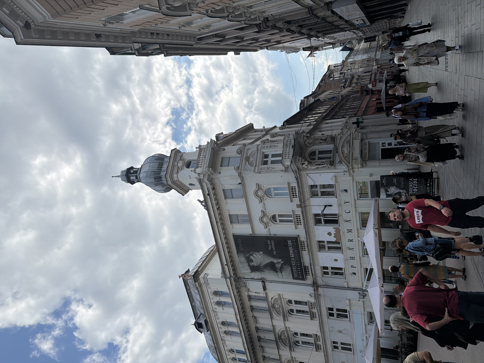 The image shows a street scene in Sarajevo, Bosnia and Herzegovina, likely in the Baščaršija area, known for its historic architecture and bustling pedestrian streets. The foreground features a group of people walking, with a man in a red t-shirt prominent on the right. Behind them, a large, ornate white building dominates the left side of the frame. A large banner displaying an image of an eye and text reading "SREBRENICA GENOCIDE MEMORIAL EXHIBITION" is prominently displayed on this building. Below this banner, a sign for "PHARMACY" is visible. The architecture of the buildings on both sides of the street is characteristic of Austro-Hungarian influence, with detailed facades, arched windows, and decorative elements. The sky above is partly cloudy with a mix of blue sky and white clouds. The street itself appears to be paved with cobblestones or similar material. Several cafes with white umbrellas are set up along the sidewalk. The overall impression is of a lively urban environment with a strong historical and cultural presence.
