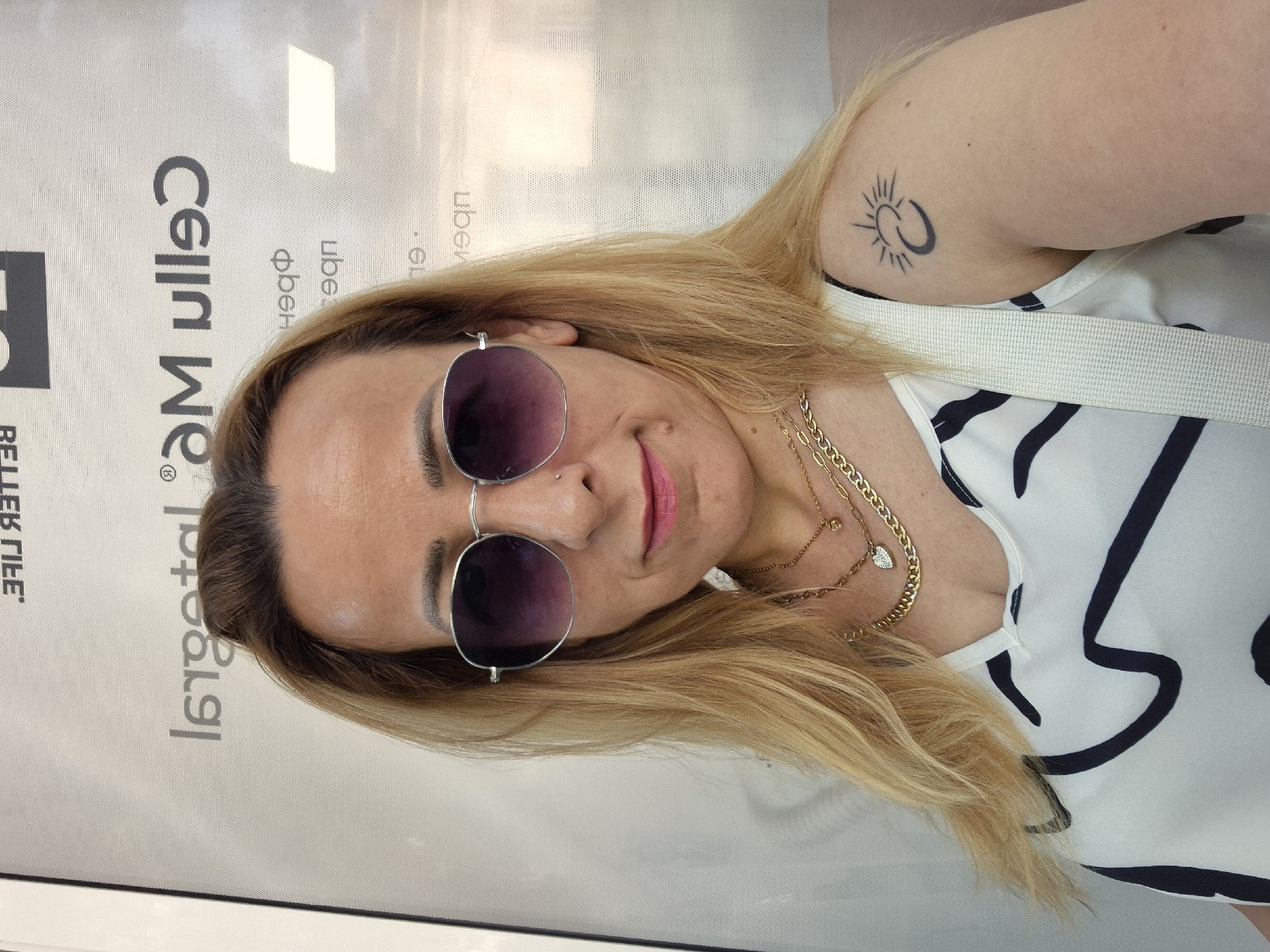A fair-skinned woman with blonde hair and sunglasses takes a selfie. The woman is wearing a white and black patterned top, with several gold necklaces, one with a heart pendant. She has a tattoo on her left shoulder of a sun and moon. The background is a textured wall with text visible.