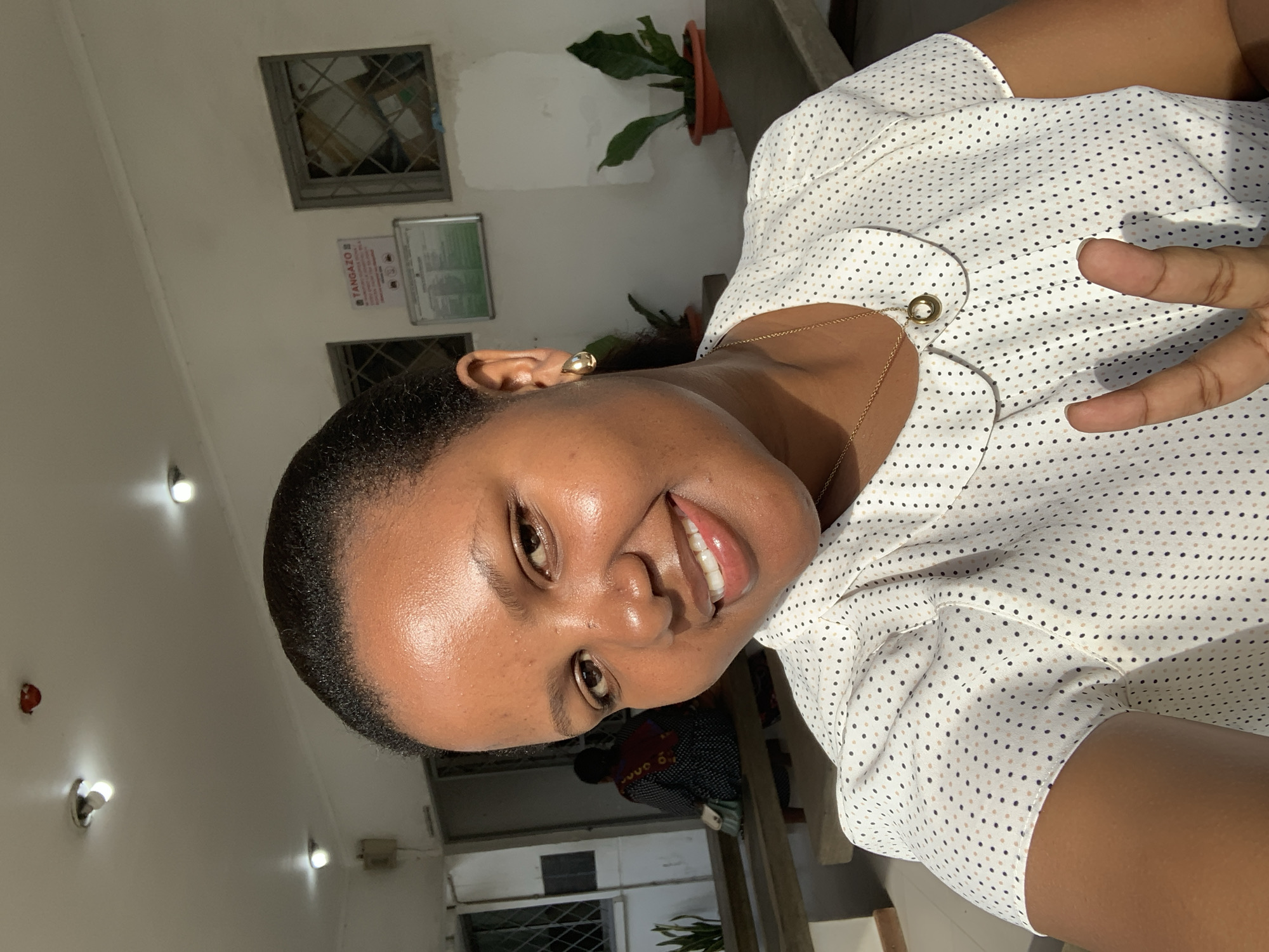 A medium shot captures a woman with dark skin and dark hair pulled back, looking directly at the camera and smiling. She is wearing a white collared shirt with small black and tan polka dots, a gold necklace with a circular pendant, and gold teardrop earrings. Her right hand is raised, with her index and middle fingers extended in a peace sign. The background appears to be outdoors, possibly a patio or courtyard area. Sunlight illuminates the scene from the right, casting shadows. In the background, there are windows with barred panes, a sign that reads "TANGAZO," two potted plants, and another person seated on a bench. The overall environment suggests a public or institutional setting within a city like Dar es Salaam, Tanzania.