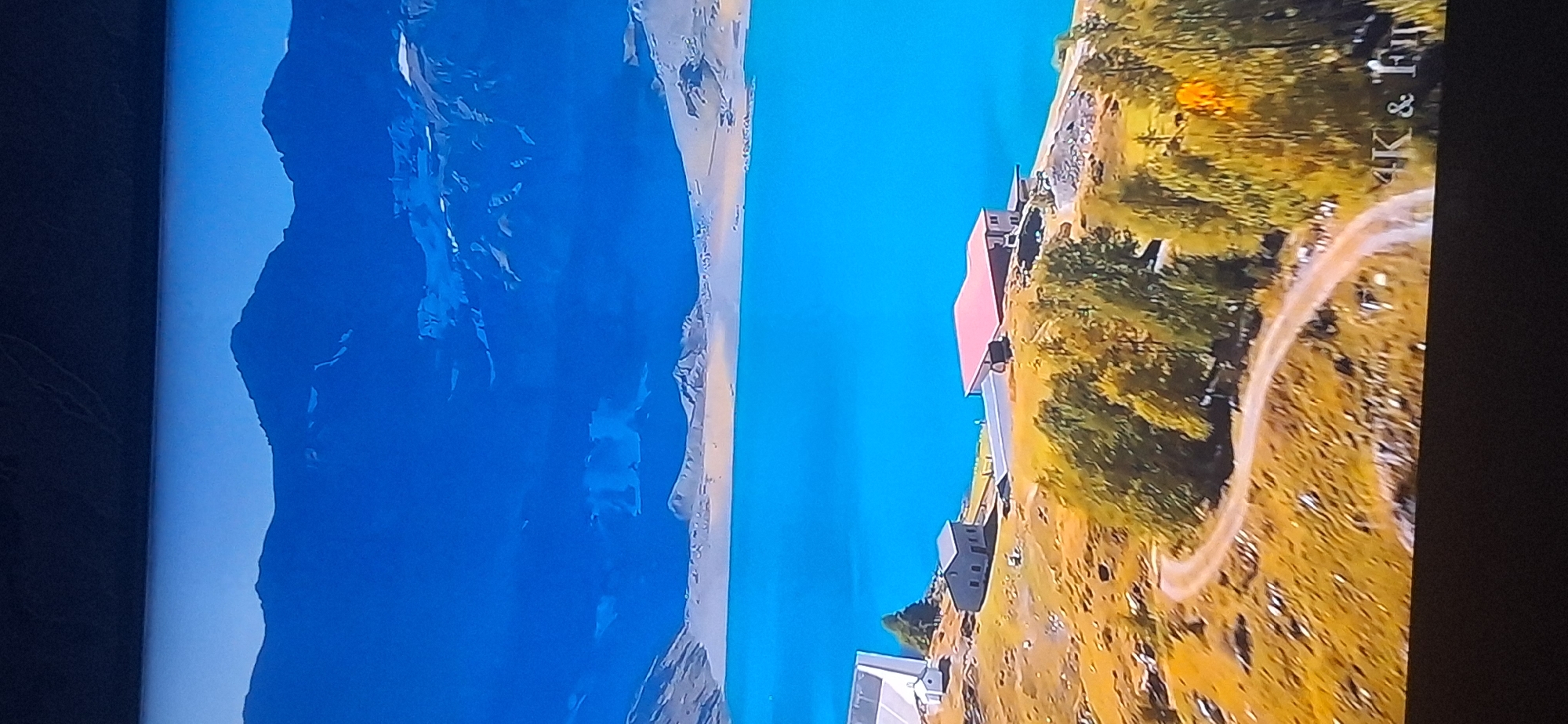The image depicts a scenic mountain landscape with a vibrant turquoise lake in the foreground. The mountains are rugged and snow-capped, suggesting a high-altitude location. On the shore of the lake, there are several buildings, including what appears to be a house with a red roof and a smaller gray building. A dirt road winds through a grassy, autumnal landscape with scattered trees. In the distance, the clear blue sky meets the imposing mountain range. The text "4K & Full" is visible in the lower right corner of the image.
There is no identifiable information within the image that places this scene in Bila Tserkva, Ukraine. The visual elements are consistent with a mountainous region, not a typical Ukrainian city.