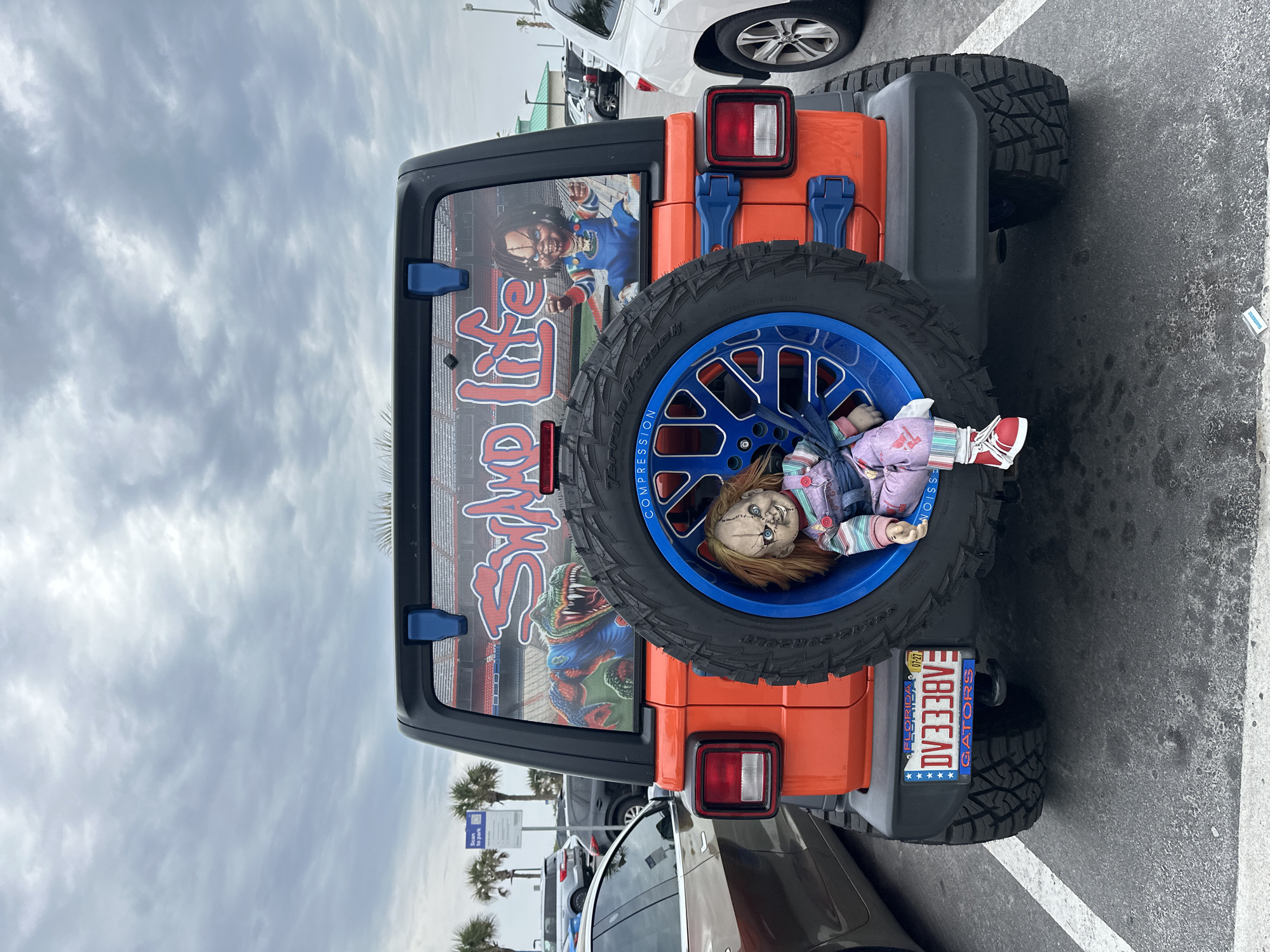 This image shows the back of an orange Jeep Wrangler, parked in a parking lot. The vehicle has custom details: a spare tire with a bright blue rim and a Chucky doll positioned within the tire. The back window has a custom graphic. A Florida license plate is visible, with the text "DV338VE." The license plate also indicates support for the Florida Gators. Cars surround the Jeep, and palm trees are visible in the background. The scene is observed to be in the city of Jacksonville Beach, United States of America.