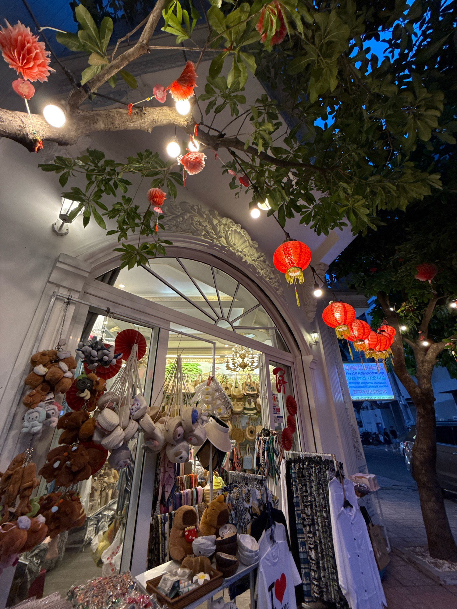 The image depicts a street view at night in Nha Trang, Vietnam, focusing on the exterior of a shop decorated for a festive occasion. Red paper lanterns are strung across the branches of a tree and along the building's awning, casting a warm glow. Several small, decorative red paper flowers are also hanging. The shop's facade features a large, arched window with glass panes, behind which a brightly lit interior is visible, showcasing an array of merchandise. In front of the window, various items are displayed on hanging racks and tables. This includes an assortment of plush toys, possibly teddy bears and other stuffed animals, as well as clothing items like t-shirts and shirts hanging on display racks. Some hats and bags are also visible among the merchandise. A street lamp is mounted on the building wall. Beyond the shop, a tree with dark green leaves is prominent, its branches intertwined with the festive decorations. The sky above is dark blue, suggesting it is nighttime. Further in the background, part of a street is visible with parked vehicles and what appears to be more buildings. A sign with Vietnamese and possibly Korean text is also present on the exterior. The overall atmosphere is festive and commercial.