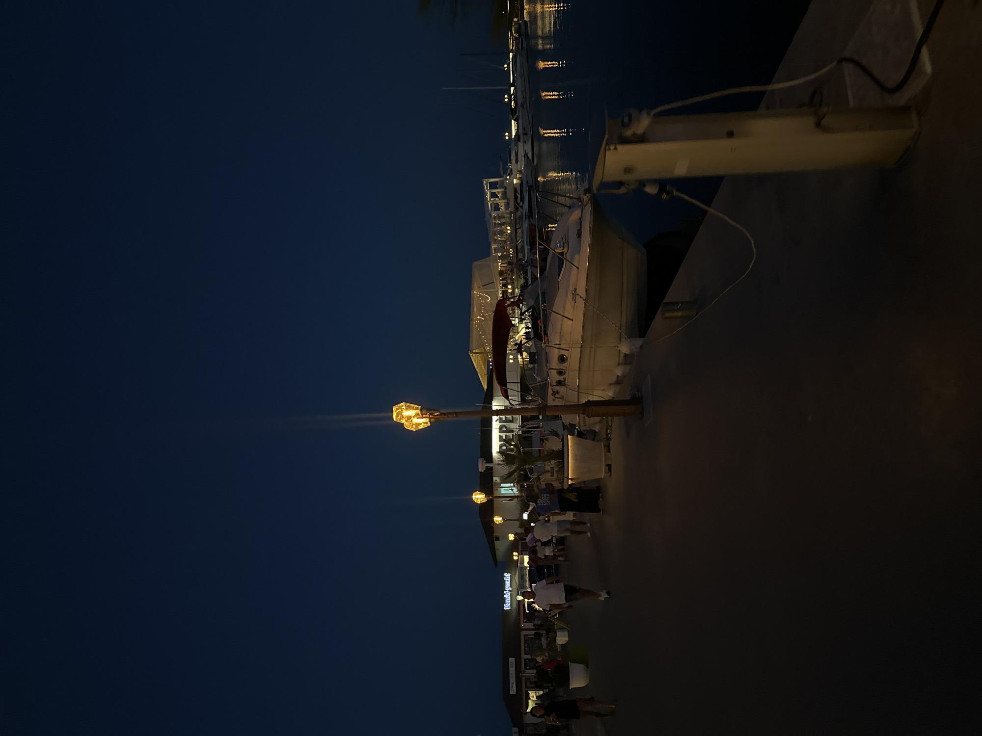 At dusk, people stroll along a paved walkway in the city of Sveti Vlas, Bulgaria, adjacent to a harbor filled with moored boats. The sky is a deep blue, indicating late evening. Several boats are visible, including a white motorboat docked near the walkway. In the background, buildings with illuminated signs, including one that reads "Vendid yacht," suggest restaurants or shops. A decorative lamppost with three glowing amber lights stands prominently on the walkway. The scene is dimly lit by the artificial lights of the buildings and the lamppost, reflecting on the calm water.