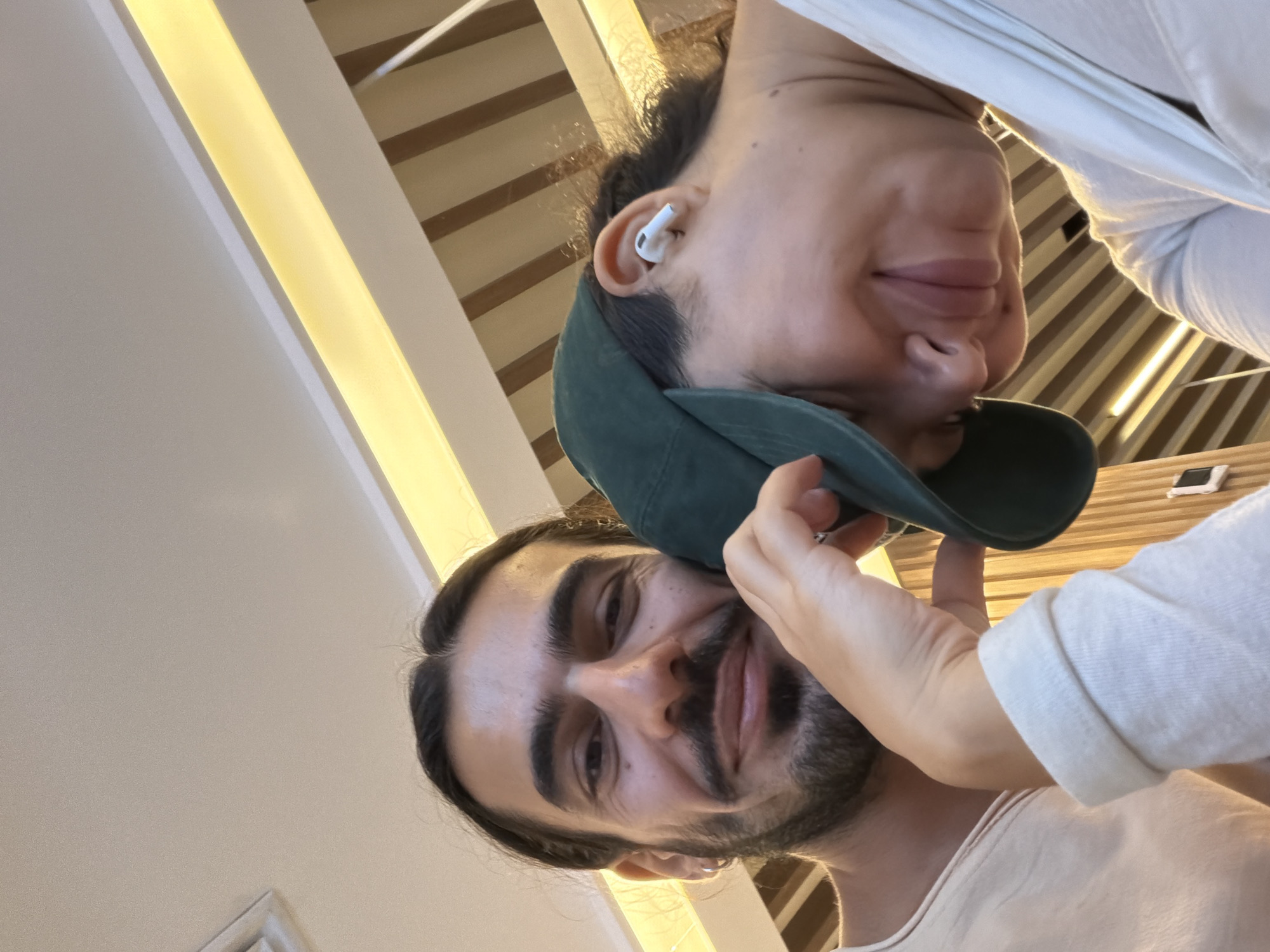 This close-up image captures two individuals in an indoor setting, likely taken as a selfie from a low angle. In the foreground, a smiling male with a beard, mustache, and dark hair tied back looks directly at the camera. He is wearing a light-colored top. Above him, and inverted in the frame, is a female, also smiling. She wears a white top and a dark green baseball cap, with a white earbud visible in her left ear. Her hand is seen holding the brim of her cap, which is resting on the male's head, suggesting a playful and affectionate interaction between the two subjects.
The background features modern interior architectural elements, including a bright, linear warm yellow light fixture mounted on a white wall or ceiling, and a ceiling with distinct wooden slats. The bright artificial lighting suggests the photo could have been taken at any time of day indoors. Both individuals express happiness and a lighthearted mood. The setting, located in North Kuta, Indonesia, appears to be a contemporary space, possibly a cafe, restaurant, or hotel lobby. No visible text is present in the image.