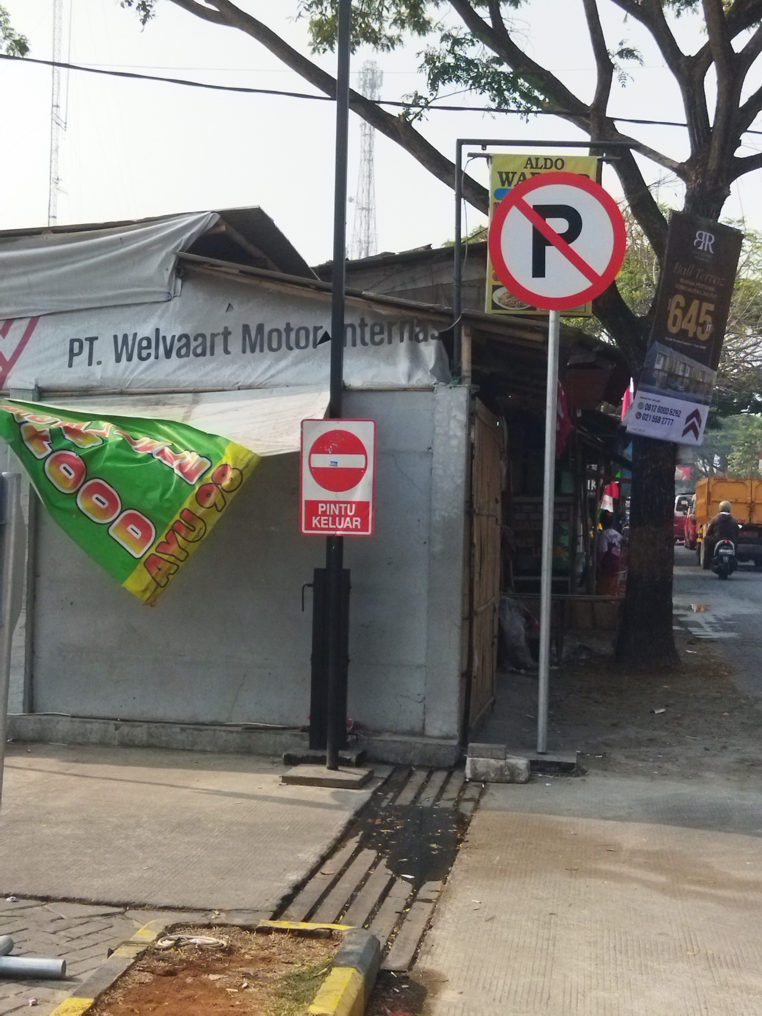 The image shows a street scene in Sepatan, Indonesia, likely during the daytime given the bright lighting. In the foreground, a paved walkway with a yellow and black striped curb is visible. To the left, a portion of a concrete wall and some rolled-up pipes are present.

Dominating the mid-ground is a stall or small building with a grey, tarp-covered roof. A large green banner with yellow lettering reading "FOOD AYU 98" is draped over the left side of the stall. The text "PT. Welvaart Motor" is printed on the grey tarp. A black pole stands in front of the stall, supporting two signs. The lower sign is red and white with the text "PINTU KELUAR" (Exit Door) and a white circle with a red horizontal bar. Above this is a yellow sign with "ALDO" and "WA" visible.

To the right of the stall, a tall tree with branches extends into the frame. Attached to the tree are several advertisements. One prominent advertisement is for "RR Bail Terras" with the number "645" displayed large. Another smaller sign below it displays phone numbers, "0812 6000 6262" and "021 568 2777".

In the background, a street runs parallel to the stall. A yellow truck is parked on the right side of the street, and a person on a black motorcycle wearing a helmet is riding away from the camera. Other vehicles and structures are faintly visible further down the street. The sky is overcast or hazy. The overall impression is of a commercial street with temporary structures and ongoing activity.