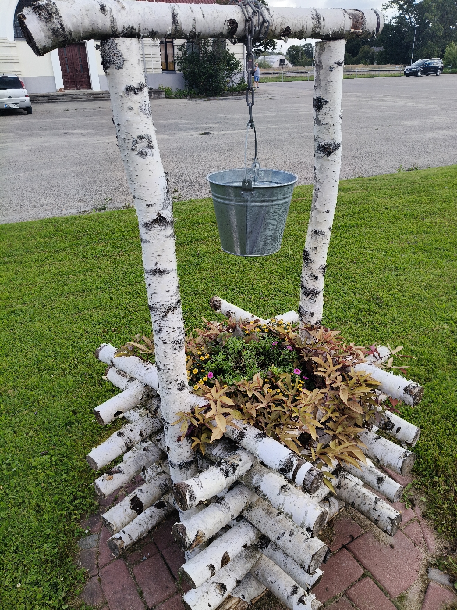 This is a medium-shot, eye-level, outdoor photograph of a decorative well structure made of birch logs. The structure is situated on a patch of green grass next to a paved area, likely a sidewalk or parking lot. The well is topped with a horizontal birch log supported by two vertical birch logs, creating an A-frame. A metal chain hangs from the horizontal log, suspending a galvanized metal bucket filled with water. Below the well's frame, the base is constructed from horizontally stacked birch logs, forming a circular enclosure. Inside this enclosure, a flower bed contains a variety of plants with reddish-brown leaves and small yellow and pink flowers. In the background, a road and a parking lot are visible, with a silver car parked on the left and a black SUV driving in the distance on the right. The sky is overcast. The scene is located in Koknese, Latvia.