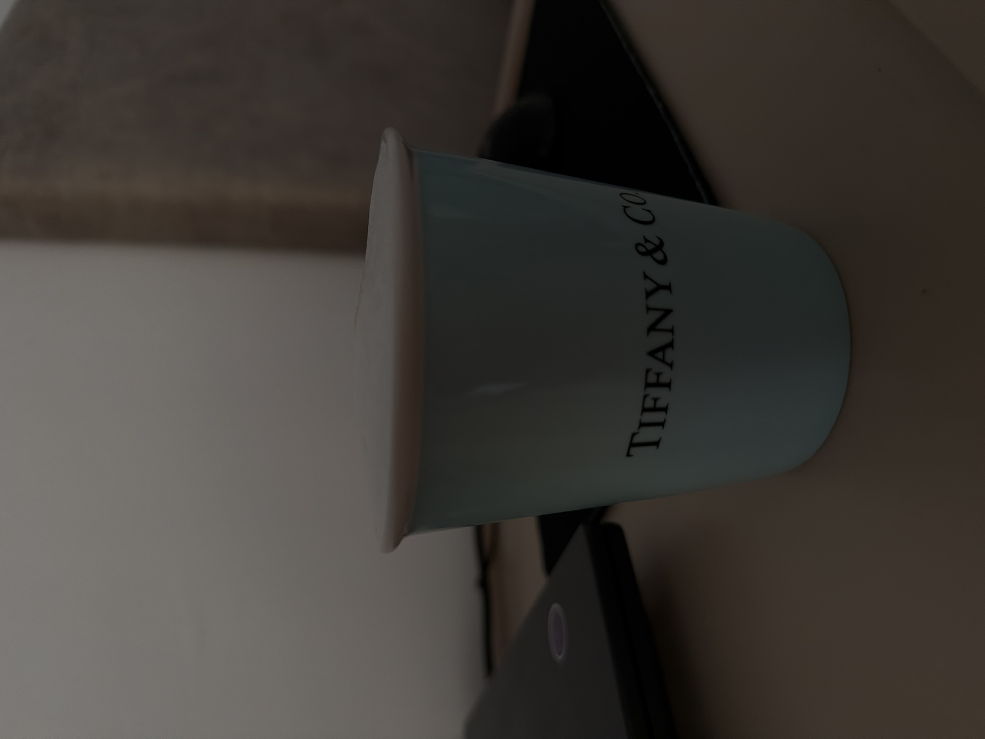 The image shows a close-up of a light blue Tiffany & Co. mug. The mug is cylindrical and has a white rim. The "TIFFANY & CO" logo is printed in black script down the side of the mug. The mug appears to be made of ceramic. It is positioned on a dark surface, possibly a table or a shelf, and part of a dark object with a circular illuminated button is visible below the mug. The background is blurred and appears to be a neutral, indoor setting with soft lighting. The image is oriented vertically.