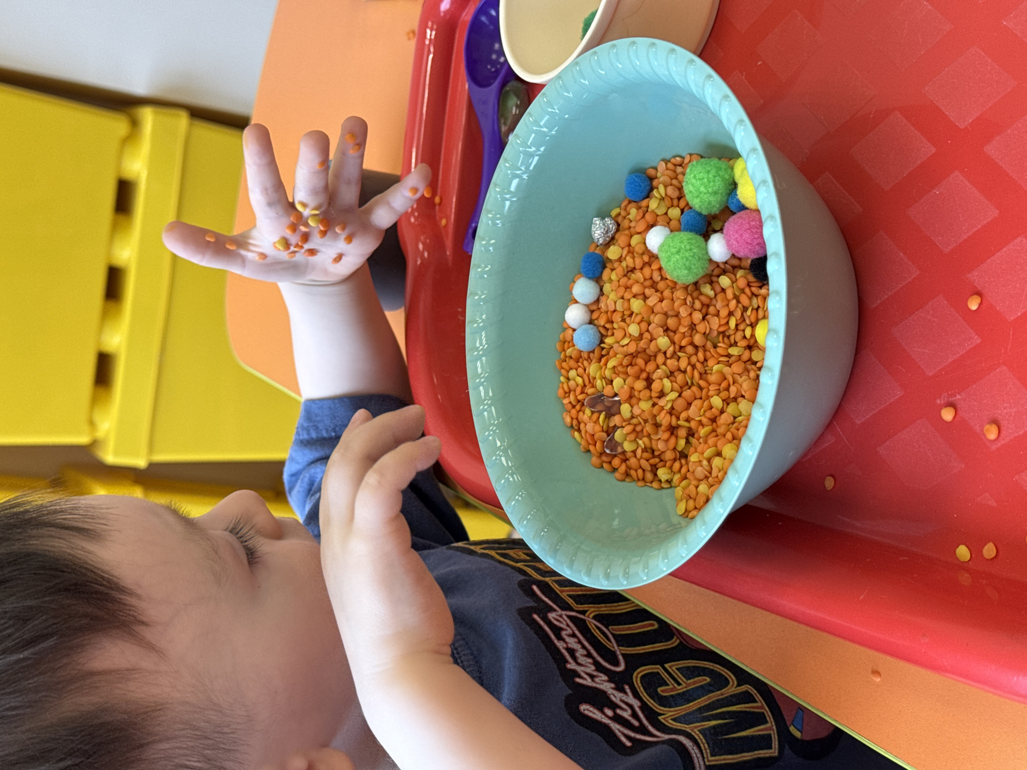 A young child is captured in an indoor setting, likely a play area or home in Izhevsk, Russia, engaging in sensory play. The child's profile is visible on the left side of the image, showing dark hair and a focused expression as they interact with materials on a bright red plastic table. Their right hand is open and held upwards, covered with numerous small orange and yellow lentils, indicating active exploration.
In the center, a light blue bowl is filled with a vibrant mix of orange and yellow lentils. Interspersed within the lentils are various colorful pom-poms in shades of blue, white, green, pink, and yellow, along with a small, crumpled piece of silver foil. The bowl rests on a textured red plastic surface, which displays a repeating diamond pattern.
In the background, a bright yellow plastic structure, possibly part of a toy or storage unit, is partially visible. Behind the blue bowl, a purple scoop and a cream-colored cup or smaller bowl can be glimpsed. The child wears a dark blue t-shirt featuring stylized black and yellow graphics, prominently displaying the word "MIGHTY." The scene is brightly lit, suggesting daytime.