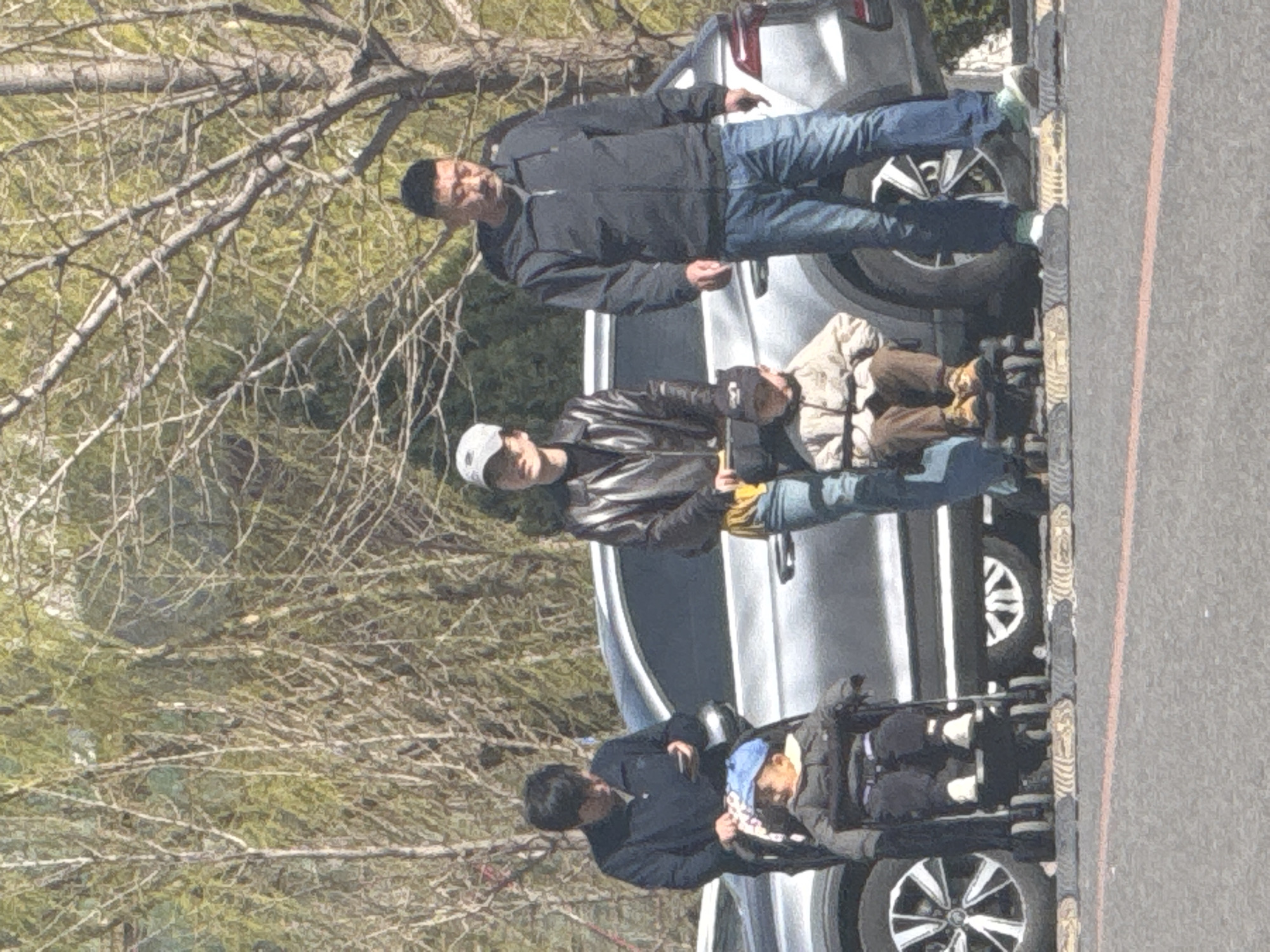 In Zhongtun, China, a family of three adults and two children are near a parked silver SUV on a sunny day. An older man in a black jacket and jeans stands by the rear tire. A younger man in a leather jacket and baseball cap holds a phone, engaging with a child sitting in a stroller. A third adult pushes a second child in a stroller. Bare trees are visible in the background.