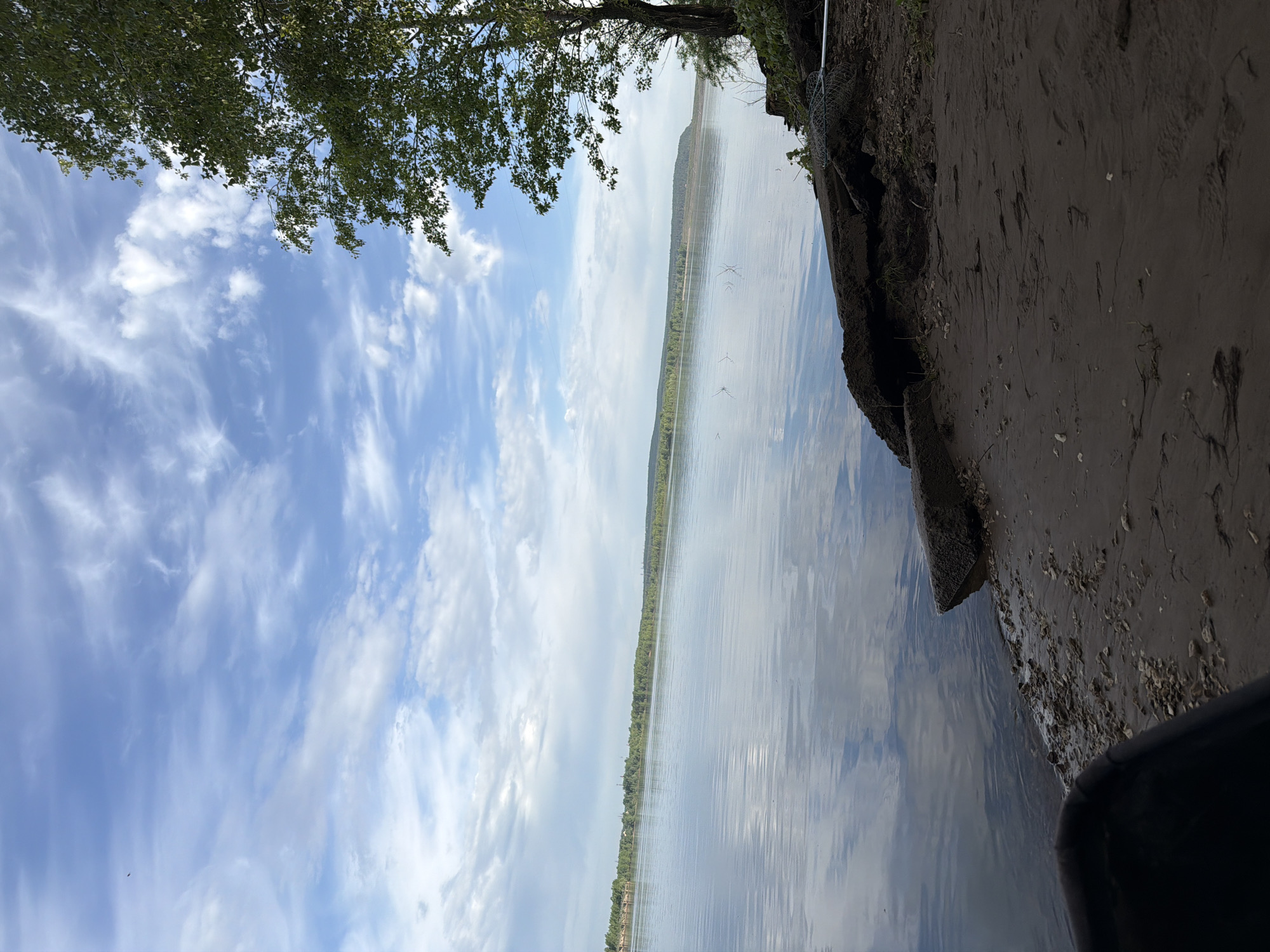The image depicts a body of water, possibly a lake or river, under a partly cloudy sky. The foreground shows a sandy shoreline on the right side. Above, trees and vegetation are present. In the center, across the body of water, is a distant shoreline with a line of trees. There are no people visible in the image. The location may be Noviy, Russia, though this cannot be verified definitively from the image alone.