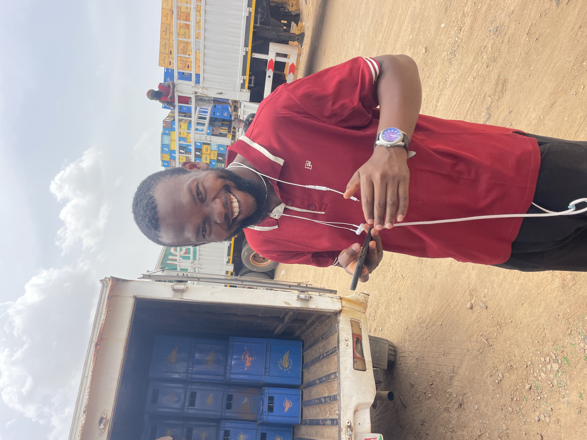 A man stands smiling in Bukuru, Nigeria, in what appears to be a transportation or delivery yard. He is wearing a maroon polo shirt, dark pants, and a watch. He holds a smartphone, with earphones dangling from his neck. Behind him is a partially opened truck, loaded with blue crates. A larger truck laden with more crates is in the background, and a person is atop it. The ground is sandy and dusty. The sky is overcast.